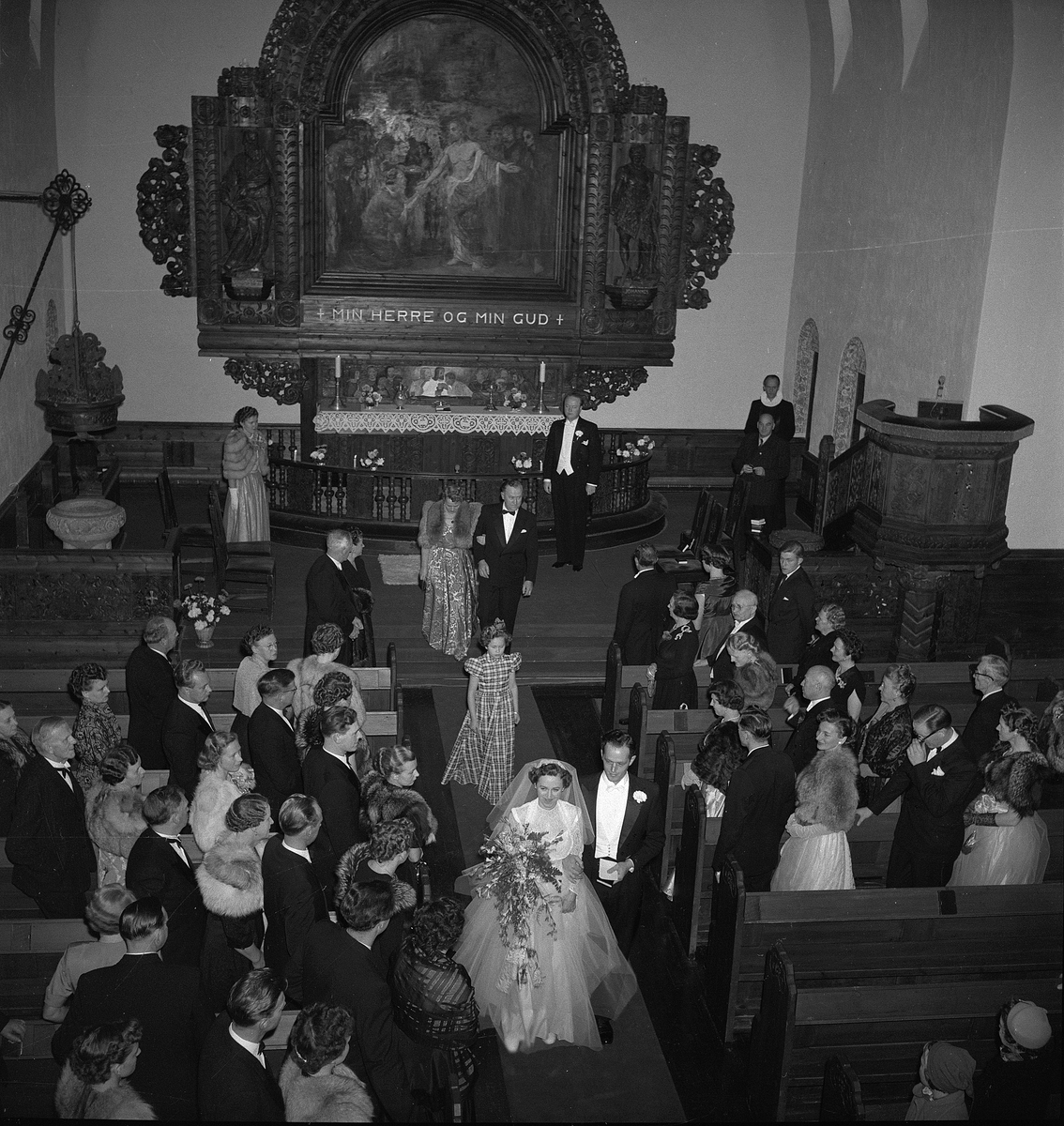 Bryllup i Rjukan kirke. Brudeparet, Walter Aasen og Hjørdis Irene Kittelsen, er på vei ut av kirken etter vielsen. Brudefølget står oppreist på hver side av midtgangen.