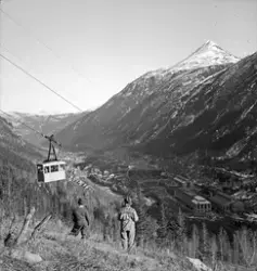 Vogn på Krossobanen med mennesker i på vei langs taubanen. Utsikt mot Rjukan fabrikker og Rjukan sentrum. To barn stående å se mot vognen.
