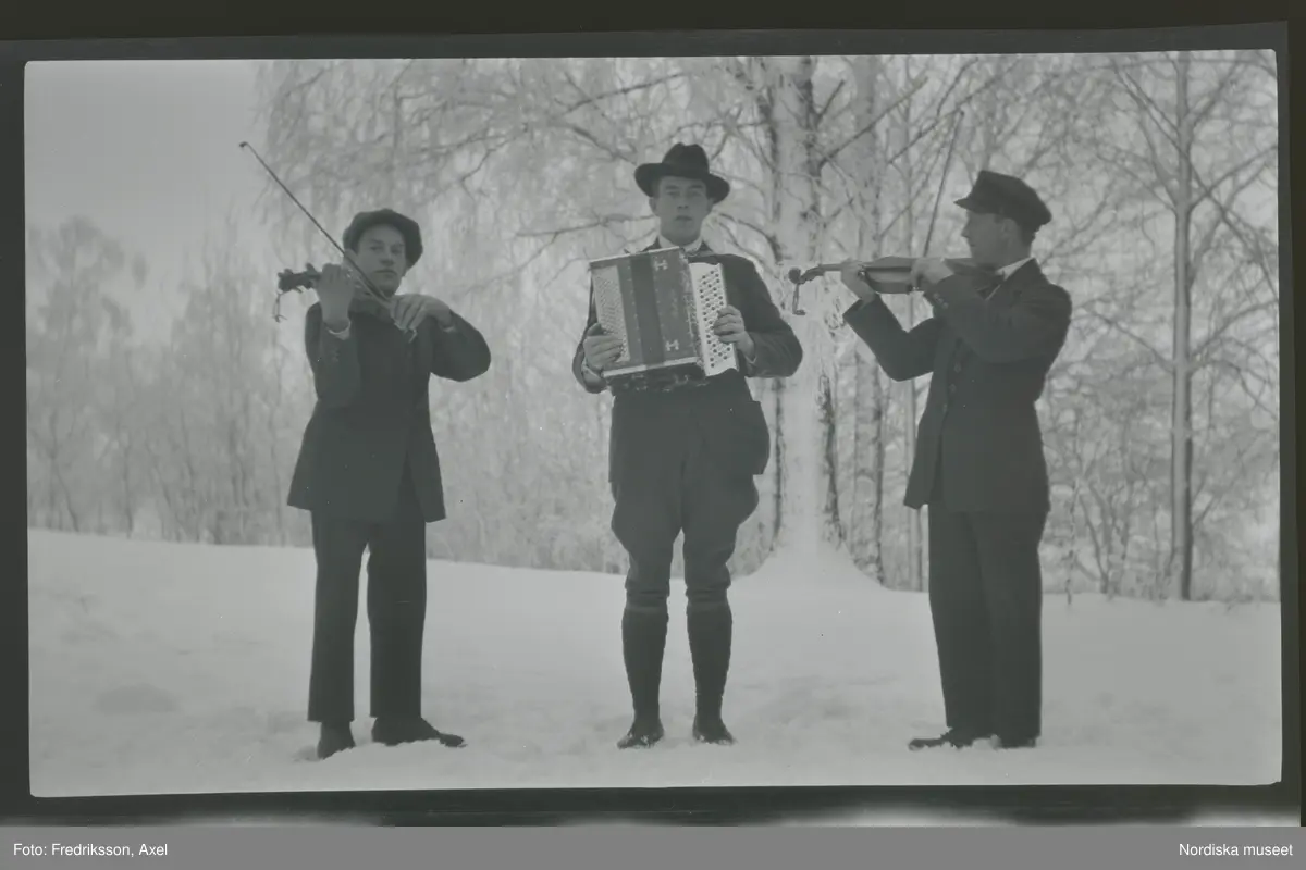 Albin, Gunnar och Johan Karlsson, Gimmersta, står utomhus i snön och spelar musik. En på dragspel och de andra två på fiol.
