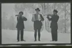 Albin, Gunnar och Johan Karlsson, Gimmersta, står utomhus i snön och spelar musik. En på dragspel och de andra två på fiol.