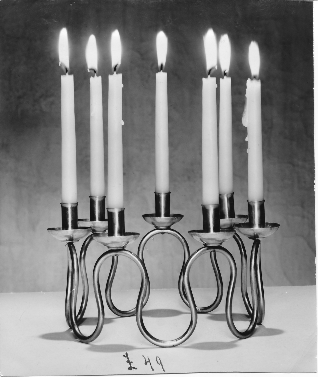 Ljusstake, "Luciakrona" för sju små stearinljus, gjord av mässingtråd som böjts i mjuk vågform. Högst upp på varje böj sitter en ljushållare fastnitad.