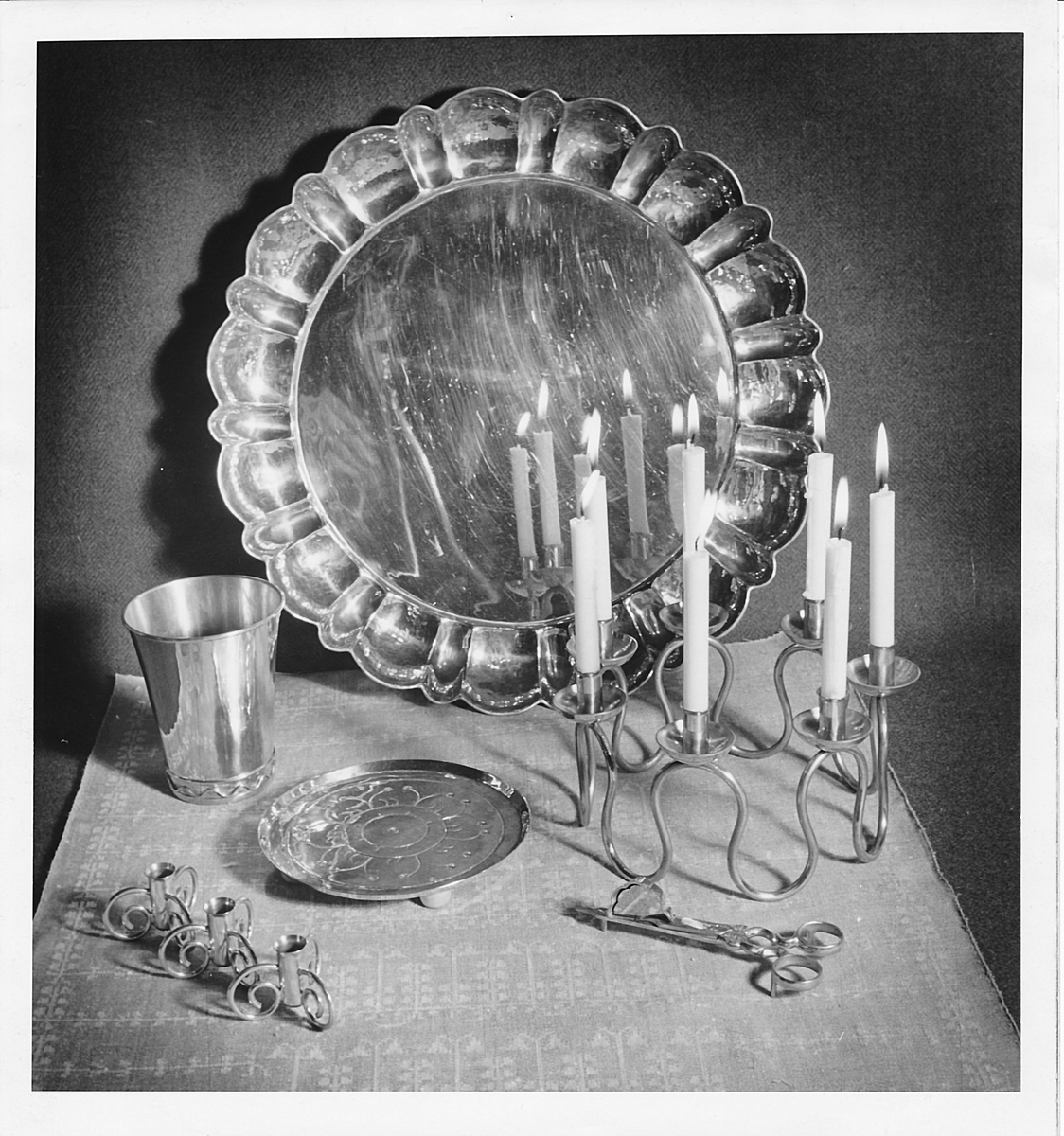 Ljusstake, "Luciakrona" för sju små stearinljus, gjord av mässingtråd som böjts i mjuk vågform. Högst upp på varje böj sitter en ljushållare fastnitad.