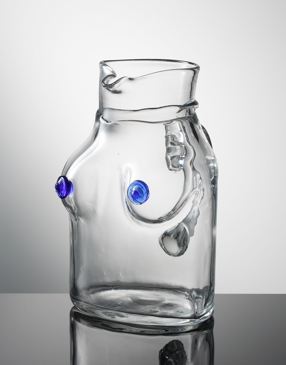 Kanna med grep, så kallad "bröstkanna", formgiven av Erik Höglund. Nedre delen i form av en kvinnlig torso med blå bröstvårtor. Halsen övergår i pip.