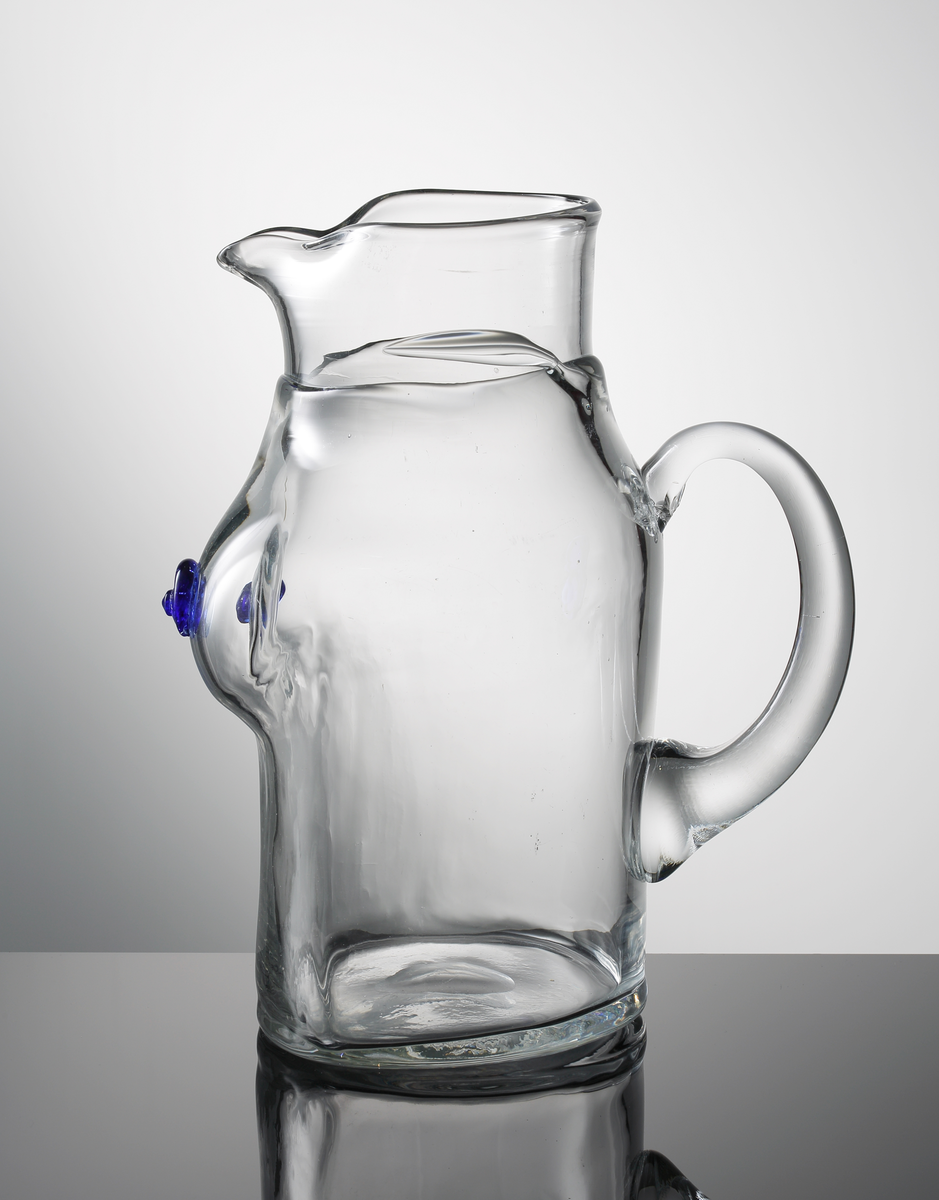 Kanna med grep, så kallad "bröstkanna", formgiven av Erik Höglund. Nedre delen i form av en kvinnlig torso med blå bröstvårtor. Halsen övergår i pip.