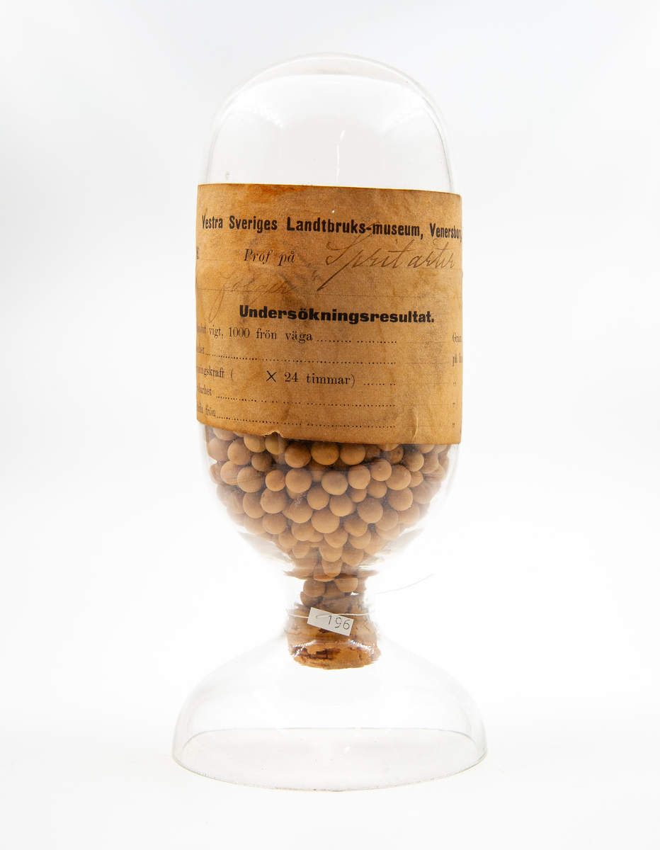Glasbehållare innehållande fröer av spritärtor (Pisum sativum var sativum).

Ingår i en frösamling under namn: ''Westra Sveriges Lantbruksmuseum''. Bildat 1880 efter lantbruksmötet i Borås. Samlingen, vilken består av glasbehållare innehållande frön från svenska kulturväxter och träd, uppställdes först på länsresidenset i Vänersborg.

Samlingen kom att bli en av Vänersborgs museums 4 grundsamlingar och uppställdes i museets övre våning vid museets öppnande 1891.

Troligen är glasbehållaren tillverkad vid Reijmyre glasbruk, ligger i Rejmyre, norr om Norrköping.
