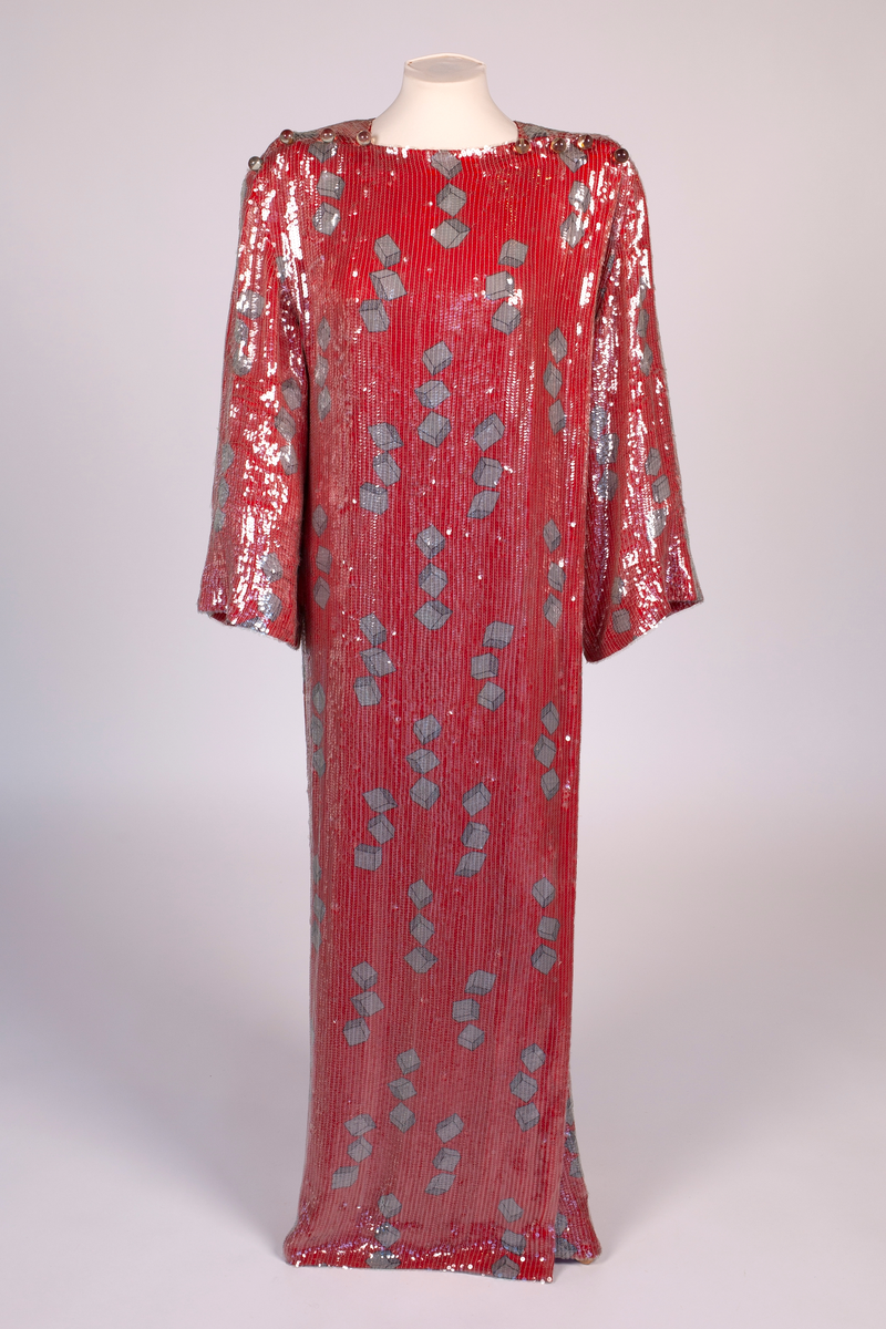 Fotsid, smal omslagskjole, kneppet på den ene skulderen og med lange vide ermer. Rødt silkestoff med påtrykt mønster i grått og svart. Mønsteret består av klosser gruppert tre og tre. Kjolen er dekket av små, gjennomsiktige plastpaljetter festet til stoffet med kjedesøm. På hver skulder er det fire store, transparente kuleknapper av plast.