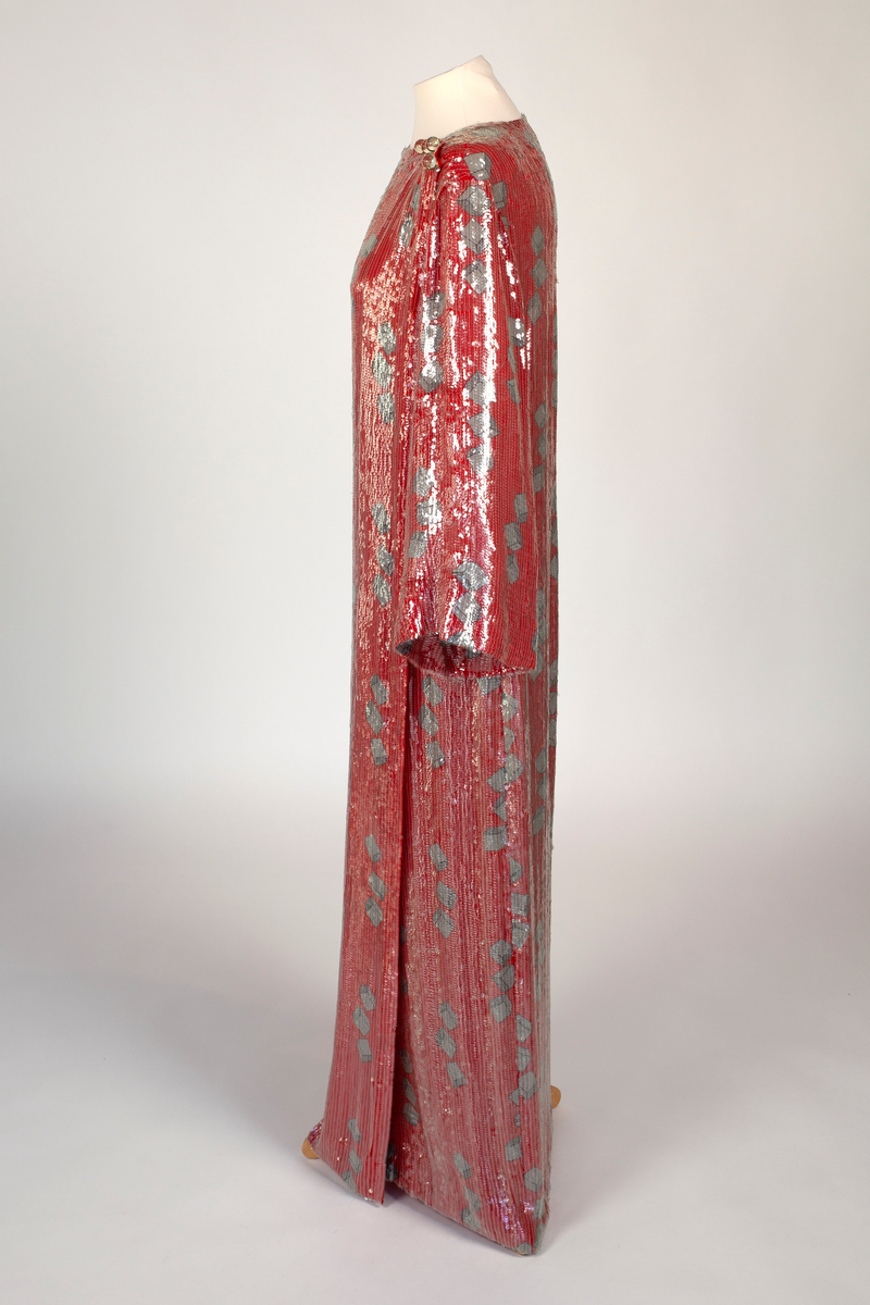 Fotsid, smal omslagskjole, kneppet på den ene skulderen og med lange vide ermer. Rødt silkestoff med påtrykt mønster i grått og svart. Mønsteret består av klosser gruppert tre og tre. Kjolen er dekket av små, gjennomsiktige plastpaljetter festet til stoffet med kjedesøm. På hver skulder er det fire store, transparente kuleknapper av plast.