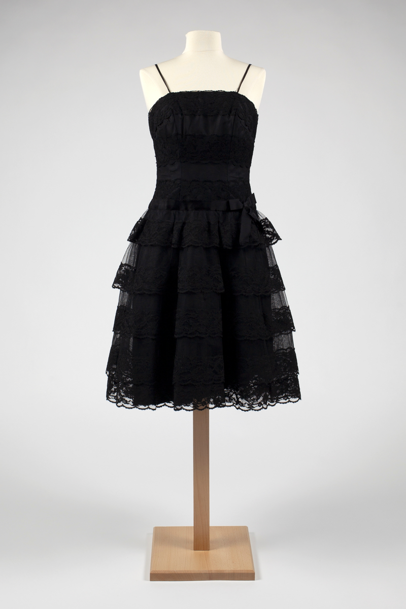 Kort, svart cocktailkjole med smale skulderstropper. Struttende skjørt med fem kapper av tarlatan (bomull) med blondekanter i avtagende lengde oppover. Under er det et enkelt tarlatansskjørt med et svart taftskjørt under, innerst en hel underkjole av grovt, appretert glanset stoff. Tilsittende liv med innfelte blonder på tarlatan, over silketaft. Glidelås bak. Forholdsvis langt liv med stivere. Svart sløyfe i venstre side i livlinjen.