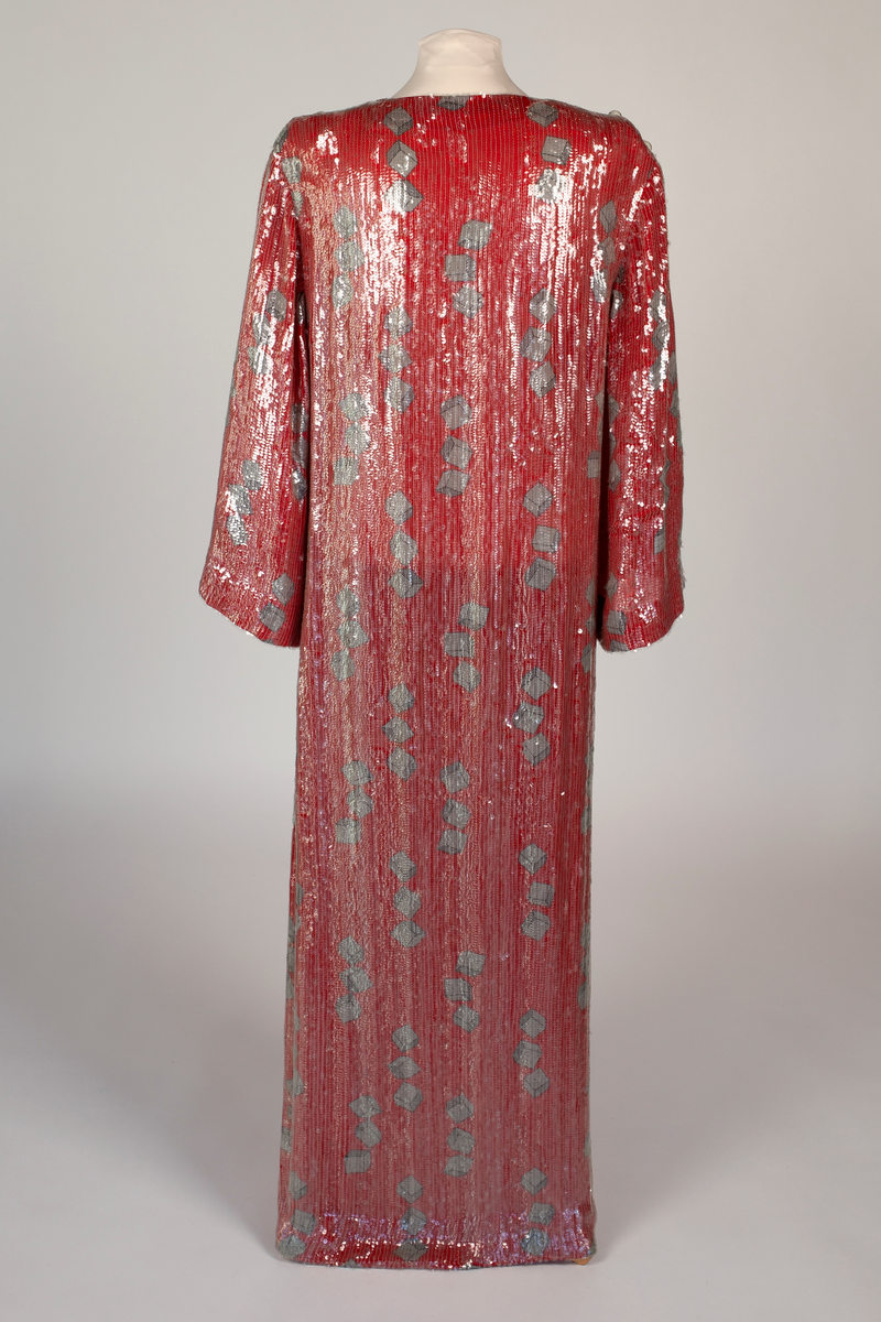 Fotsid, smal omslagskjole, kneppet på den ene skulderen og med lange vide ermer. Rødt silkestoff med påtrykt mønster i grått og svart. Mønsteret består av klosser gruppert tre og tre. Kjolen er dekket av små, gjennomsiktige plastpaljetter festet til stoffet med kjedesøm. På hver skulder er det fire store, transparente kuleknapper av plast.