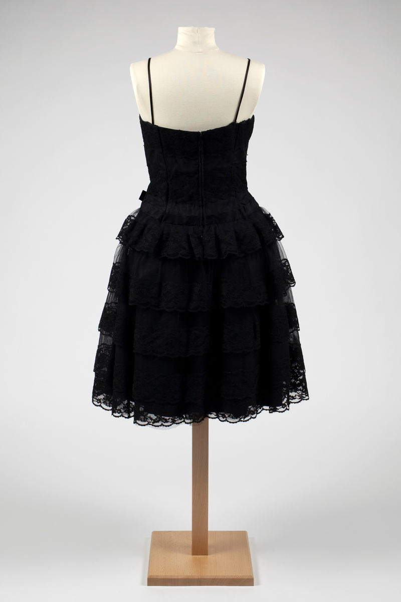 Kort, svart cocktailkjole med smale skulderstropper. Struttende skjørt med fem kapper av tarlatan (bomull) med blondekanter i avtagende lengde oppover. Under er det et enkelt tarlatansskjørt med et svart taftskjørt under, innerst en hel underkjole av grovt, appretert glanset stoff. Tilsittende liv med innfelte blonder på tarlatan, over silketaft. Glidelås bak. Forholdsvis langt liv med stivere. Svart sløyfe i venstre side i livlinjen.