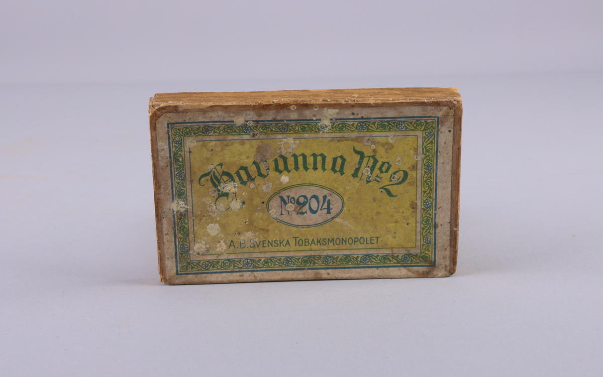 Lock till ask, av hårdpapp som utvändigt klätts i trämönstrat papper. På locket, gul etikett med grönt tryck: "Havanna no 2, no 204 AB Svenska Tobaksmonopolet".