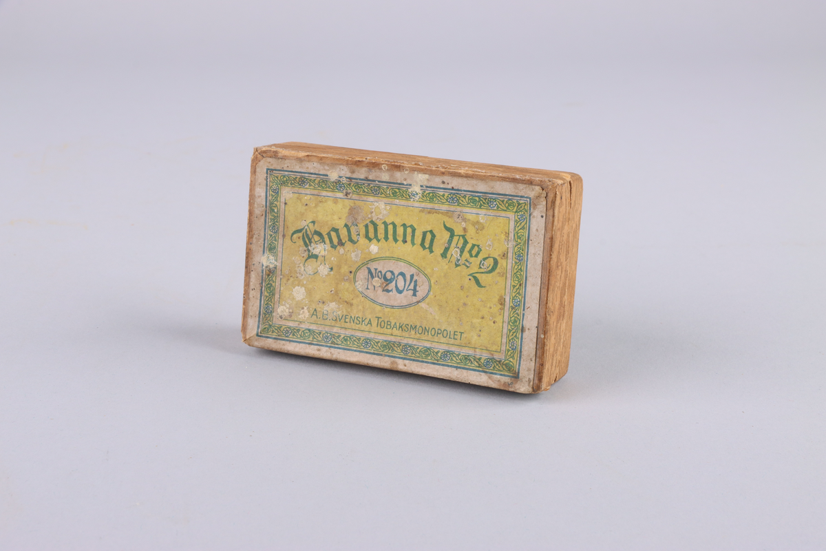 Ask, i två delar, av hårdpapp som utvändigt klätts i trämönstrat papper. På locket, gul etikett med grönt tryck: "Havanna no 2, no 204 AB Svenska Tobaksmonopolet". 