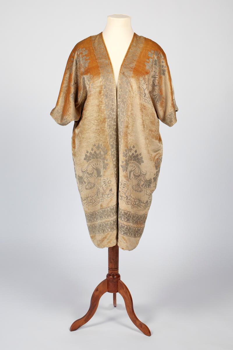 Knelang jakke av tynn, myk, aprikosfarget silkefløyel, med påtrykt mønter i sølv. Helfôret med grå, ripsartet silke. Jakken er åpen foran, med rettvinklede ermer, og er sterkt kimonoinspirert. Ermene er albulange og skåret iett med henholdsvis forstykkene og ryggstykket. For- og bakstykker er sammensydd i sidene, på skulderen og langs ermenes overside. Ryggen er helskåret, med relativ dyp V-ringning i nakken. Jakken er rikt dekorert med sølvfarget trykk av vridde treformer, blader og blomster. Hovedmotivet er plassert på hver hofte foran, og gjentatt på brystpartiet. På ryggen er dette tilsvarende plassert, det hele innrammet av en bord med stiliserte vinranker. Borden er dobbel i nederkant, men den øverste av disse bordene føres parallelt opp langs ryggens midte, fordeler seg i nakken og føres ned langs åpningen midt foran.
