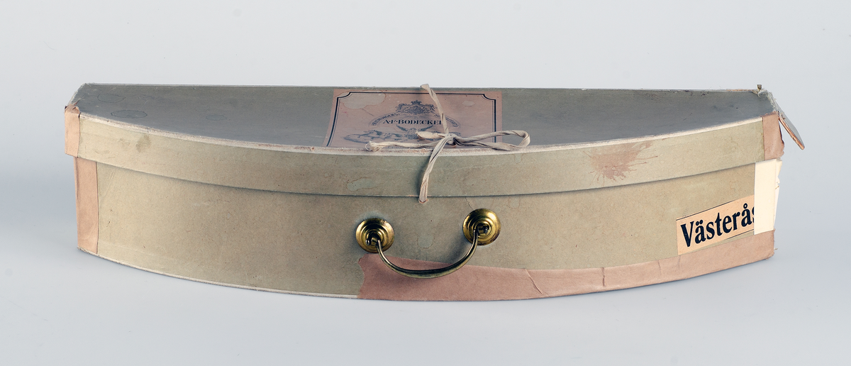 Kartong för förvaring av uniformsmössa UM07962e.  Brungrön kartong med metallhandtag. Locket hålls ihop med ett band. På ena sidan märkt med etikett H.M. Konungens Hofleverentör. A.F. Bodecker. Fabriksmärke. 6 fredgatan 6. STOCKHOLM. I mitten av etiketten staty med vingar och hästar i tyglar. Vidare på sidan en etikett för transport märkt VÄSTERÅS Fr. Stockholm C.  5767.