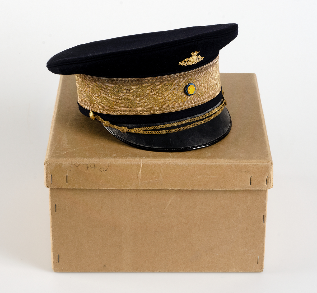 Kartong för förvaring av uniformsmössa UM07962d. Brun kartong med lock. På locket en rund etikett med texten Militärekiperings AB. MEA Norrmalmstorg Stockholm