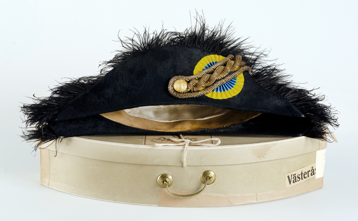 Kartong för förvaring av uniformsmössa UM07962e.  Brungrön kartong med metallhandtag. Locket hålls ihop med ett band. På ena sidan märkt med etikett H.M. Konungens Hofleverentör. A.F. Bodecker. Fabriksmärke. 6 fredgatan 6. STOCKHOLM. I mitten av etiketten staty med vingar och hästar i tyglar. Vidare på sidan en etikett för transport märkt VÄSTERÅS Fr. Stockholm C.  5767.