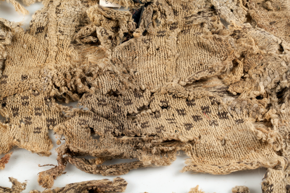 Fragment av stickad tröja med mönsterstickad bård i brunt och beige ullgarn. Bålen i 1-trådigt ullgarn mönsterstickad i rutmönster i brunt, natursvart och beige. Ärmarna är rundstickade i 2-trådigt ullgarn i naturvit, natursvart, brunt och beige. Sprund mitt fram 10 cm långt. Halsurringning och sprund mitt fram kantad med samma tyg som finns som lagningar på UM23252 och A 38 (UM ?). Hyska och hake i mässing mitt fram.