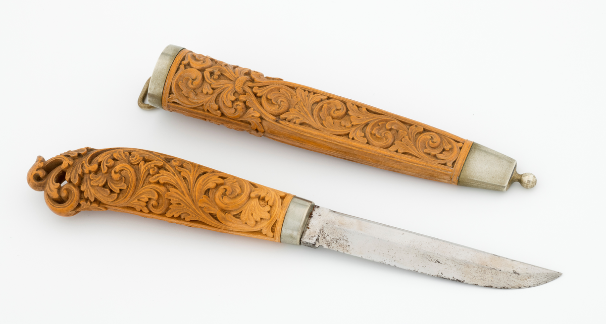 Tollekniv i slire laget av den kjente treskjæreren Ole Olsen Moene (1839-1908) fra Oppdal i Sør-Trøndelag. Kniven har blad som antas å være av jern og stål, mens skaftet og slira er utført i tre, men med beslag av nysølv. 

Kniven er 19,3 centimeter lang. Bladet utgjør 8,9 centimeter av dette lengdemålet. Det er 14,9 millimeter bredt bakerst ved brystningen. Her er tjukkelsen 3,3 millimeter. Bladryggen er noenlunde rettlinjet, men særlig den bakre delen er avfaset mot sidene. Slipefasene er opptil 9 millimeter brede. Bladet er blankpolert. Det har ingen stempler eller andre signaturer som tydeliggjør produsenten, men mye tyder vel på at dette er et «fabrikkblad» - altså en masseprodusert komponent.

Tangen i den bakre enden av skaftet er tappet inn i et snaut 10,5 centimeter langt skaft, lagd av det for nordmenn eksotiske treslaget buksbom (Buxus sempervirens). Dette er en buskvekst som har sin opprinnelige utbredelse i det submeriterrane området. Den fremre skaftenden er forsterket med en 7 millimeter bred skaftholk med glatt overflate. Skaftet er konvekst i formen, og det har, som på knivskaft flest, et ovalt tverrsnitt. Den bakre skaftenden er en aning krum - mot buksida. Skaftet har flatedekkende utskåret dekor, elegant formete akantusranker i cirka 2 millimeter høyt relieff, organisert på to flater, ei på hver av skaftets «bredsider». I den bakre enden er akantusbladene gjennombrudte. 

Slira er et 14,4 centimeter langt hylster, sammensatt av to buete buksbomplater med buete tverrsnitt, holdt ihop av en 5,7 millimeter bred munningsholk i den øvre enden og en 2,6 centimeter høy doppsko med endeknopp i den nedre enden. Bøde doppskoen og munningsholken har glatte overflater. De to buksbomskiene har flatedekkende akantusdekor. På de delene som skal omslutte bladet er akantusrankene innrammet av to ribber som møtes cirka 3,5 centimeter under sliremunningen. Fra disse punktene voksder det rosetter som omgir den nedre (fremre) enden av knivskaftet. På slira er akantusrelieffene om lag 1 millimeter høye. På baksida av munningsholken er detr påloddet ei hempe for et oppheng, som antakelig har vært av lær. Dette opphenget mangler.

Verken kniven eller slira har signaturer som refererer til produsenten.