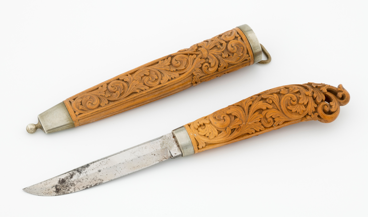 Tollekniv i slire laget av den kjente treskjæreren Ole Olsen Moene (1839-1908) fra Oppdal i Sør-Trøndelag. Kniven har blad som antas å være av jern og stål, mens skaftet og slira er utført i tre, men med beslag av nysølv. 

Kniven er 19,3 centimeter lang. Bladet utgjør 8,9 centimeter av dette lengdemålet. Det er 14,9 millimeter bredt bakerst ved brystningen. Her er tjukkelsen 3,3 millimeter. Bladryggen er noenlunde rettlinjet, men særlig den bakre delen er avfaset mot sidene. Slipefasene er opptil 9 millimeter brede. Bladet er blankpolert. Det har ingen stempler eller andre signaturer som tydeliggjør produsenten, men mye tyder vel på at dette er et «fabrikkblad» - altså en masseprodusert komponent.

Tangen i den bakre enden av skaftet er tappet inn i et snaut 10,5 centimeter langt skaft, lagd av det for nordmenn eksotiske treslaget buksbom (Buxus sempervirens). Dette er en buskvekst som har sin opprinnelige utbredelse i det submeriterrane området. Den fremre skaftenden er forsterket med en 7 millimeter bred skaftholk med glatt overflate. Skaftet er konvekst i formen, og det har, som på knivskaft flest, et ovalt tverrsnitt. Den bakre skaftenden er en aning krum - mot buksida. Skaftet har flatedekkende utskåret dekor, elegant formete akantusranker i cirka 2 millimeter høyt relieff, organisert på to flater, ei på hver av skaftets «bredsider». I den bakre enden er akantusbladene gjennombrudte. 

Slira er et 14,4 centimeter langt hylster, sammensatt av to buete buksbomplater med buete tverrsnitt, holdt ihop av en 5,7 millimeter bred munningsholk i den øvre enden og en 2,6 centimeter høy doppsko med endeknopp i den nedre enden. Bøde doppskoen og munningsholken har glatte overflater. De to buksbomskiene har flatedekkende akantusdekor. På de delene som skal omslutte bladet er akantusrankene innrammet av to ribber som møtes cirka 3,5 centimeter under sliremunningen. Fra disse punktene voksder det rosetter som omgir den nedre (fremre) enden av knivskaftet. På slira er akantusrelieffene om lag 1 millimeter høye. På baksida av munningsholken er detr påloddet ei hempe for et oppheng, som antakelig har vært av lær. Dette opphenget mangler.

Verken kniven eller slira har signaturer som refererer til produsenten.