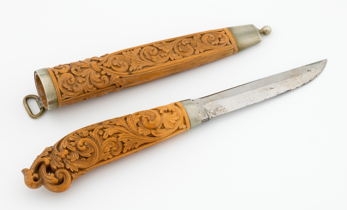 Tollekniv i slire laget av den kjente treskjæreren Ole Olsen Moene (1839-1908) fra Oppdal i Sør-Trøndelag. Kniven har blad som antas å være av jern og stål, mens skaftet og slira er utført i tre, men med beslag av nysølv. 

Kniven er 19,3 centimeter lang. Bladet utgjør 8,9 centimeter av dette lengdemålet. Det er 14,9 millimeter bredt bakerst ved brystningen. Her er tjukkelsen 3,3 millimeter. Bladryggen er noenlunde rettlinjet, men særlig den bakre delen er avfaset mot sidene. Slipefasene er opptil 9 millimeter brede. Bladet er blankpolert. Det har ingen stempler eller andre signaturer som tydeliggjør produsenten, men mye tyder vel på at dette er et «fabrikkblad» - altså en masseprodusert komponent.

Tangen i den bakre enden av skaftet er tappet inn i et snaut 10,5 centimeter langt skaft, lagd av det for nordmenn eksotiske treslaget buksbom (Buxus sempervirens). Dette er en buskvekst som har sin opprinnelige utbredelse i det submeriterrane området. Den fremre skaftenden er forsterket med en 7 millimeter bred skaftholk med glatt overflate. Skaftet er konvekst i formen, og det har, som på knivskaft flest, et ovalt tverrsnitt. Den bakre skaftenden er en aning krum - mot buksida. Skaftet har flatedekkende utskåret dekor, elegant formete akantusranker i cirka 2 millimeter høyt relieff, organisert på to flater, ei på hver av skaftets «bredsider». I den bakre enden er akantusbladene gjennombrudte. 

Slira er et 14,4 centimeter langt hylster, sammensatt av to buete buksbomplater med buete tverrsnitt, holdt ihop av en 5,7 millimeter bred munningsholk i den øvre enden og en 2,6 centimeter høy doppsko med endeknopp i den nedre enden. Bøde doppskoen og munningsholken har glatte overflater. De to buksbomskiene har flatedekkende akantusdekor. På de delene som skal omslutte bladet er akantusrankene innrammet av to ribber som møtes cirka 3,5 centimeter under sliremunningen. Fra disse punktene voksder det rosetter som omgir den nedre (fremre) enden av knivskaftet. På slira er akantusrelieffene om lag 1 millimeter høye. På baksida av munningsholken er detr påloddet ei hempe for et oppheng, som antakelig har vært av lær. Dette opphenget mangler.

Verken kniven eller slira har signaturer som refererer til produsenten.