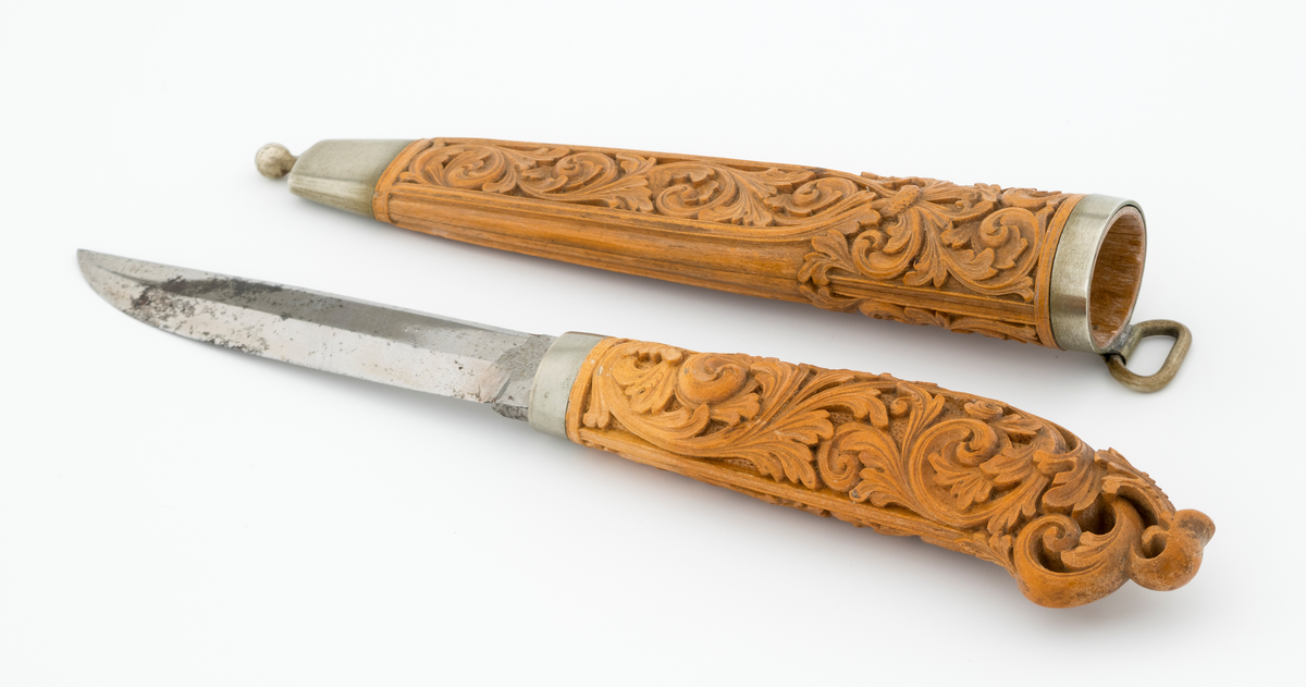Tollekniv i slire laget av den kjente treskjæreren Ole Olsen Moene (1839-1908) fra Oppdal i Sør-Trøndelag. Kniven har blad som antas å være av jern og stål, mens skaftet og slira er utført i tre, men med beslag av nysølv. 

Kniven er 19,3 centimeter lang. Bladet utgjør 8,9 centimeter av dette lengdemålet. Det er 14,9 millimeter bredt bakerst ved brystningen. Her er tjukkelsen 3,3 millimeter. Bladryggen er noenlunde rettlinjet, men særlig den bakre delen er avfaset mot sidene. Slipefasene er opptil 9 millimeter brede. Bladet er blankpolert. Det har ingen stempler eller andre signaturer som tydeliggjør produsenten, men mye tyder vel på at dette er et «fabrikkblad» - altså en masseprodusert komponent.

Tangen i den bakre enden av skaftet er tappet inn i et snaut 10,5 centimeter langt skaft, lagd av det for nordmenn eksotiske treslaget buksbom (Buxus sempervirens). Dette er en buskvekst som har sin opprinnelige utbredelse i det submeriterrane området. Den fremre skaftenden er forsterket med en 7 millimeter bred skaftholk med glatt overflate. Skaftet er konvekst i formen, og det har, som på knivskaft flest, et ovalt tverrsnitt. Den bakre skaftenden er en aning krum - mot buksida. Skaftet har flatedekkende utskåret dekor, elegant formete akantusranker i cirka 2 millimeter høyt relieff, organisert på to flater, ei på hver av skaftets «bredsider». I den bakre enden er akantusbladene gjennombrudte. 

Slira er et 14,4 centimeter langt hylster, sammensatt av to buete buksbomplater med buete tverrsnitt, holdt ihop av en 5,7 millimeter bred munningsholk i den øvre enden og en 2,6 centimeter høy doppsko med endeknopp i den nedre enden. Bøde doppskoen og munningsholken har glatte overflater. De to buksbomskiene har flatedekkende akantusdekor. På de delene som skal omslutte bladet er akantusrankene innrammet av to ribber som møtes cirka 3,5 centimeter under sliremunningen. Fra disse punktene voksder det rosetter som omgir den nedre (fremre) enden av knivskaftet. På slira er akantusrelieffene om lag 1 millimeter høye. På baksida av munningsholken er detr påloddet ei hempe for et oppheng, som antakelig har vært av lær. Dette opphenget mangler.

Verken kniven eller slira har signaturer som refererer til produsenten.