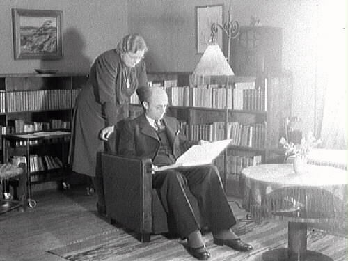 Museichef Albert Sandklef med frun Karin Sandklef i bostadens bokhörna. Albert läser och Karin står bakom honom. Fotografiet är taget i Artillerikasernen på Fästningen.