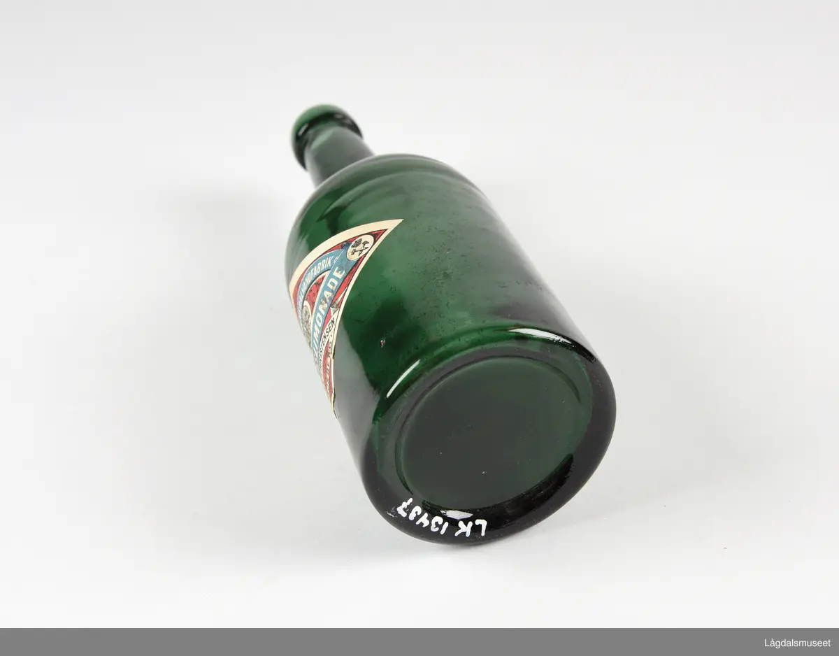 Grønn glassflaske med etikett. Etiketten har Kongsbergs gamle kommunevåpen og bergmannssymbolet korslagt hammer og bergsjern. Det siste med krone over. 