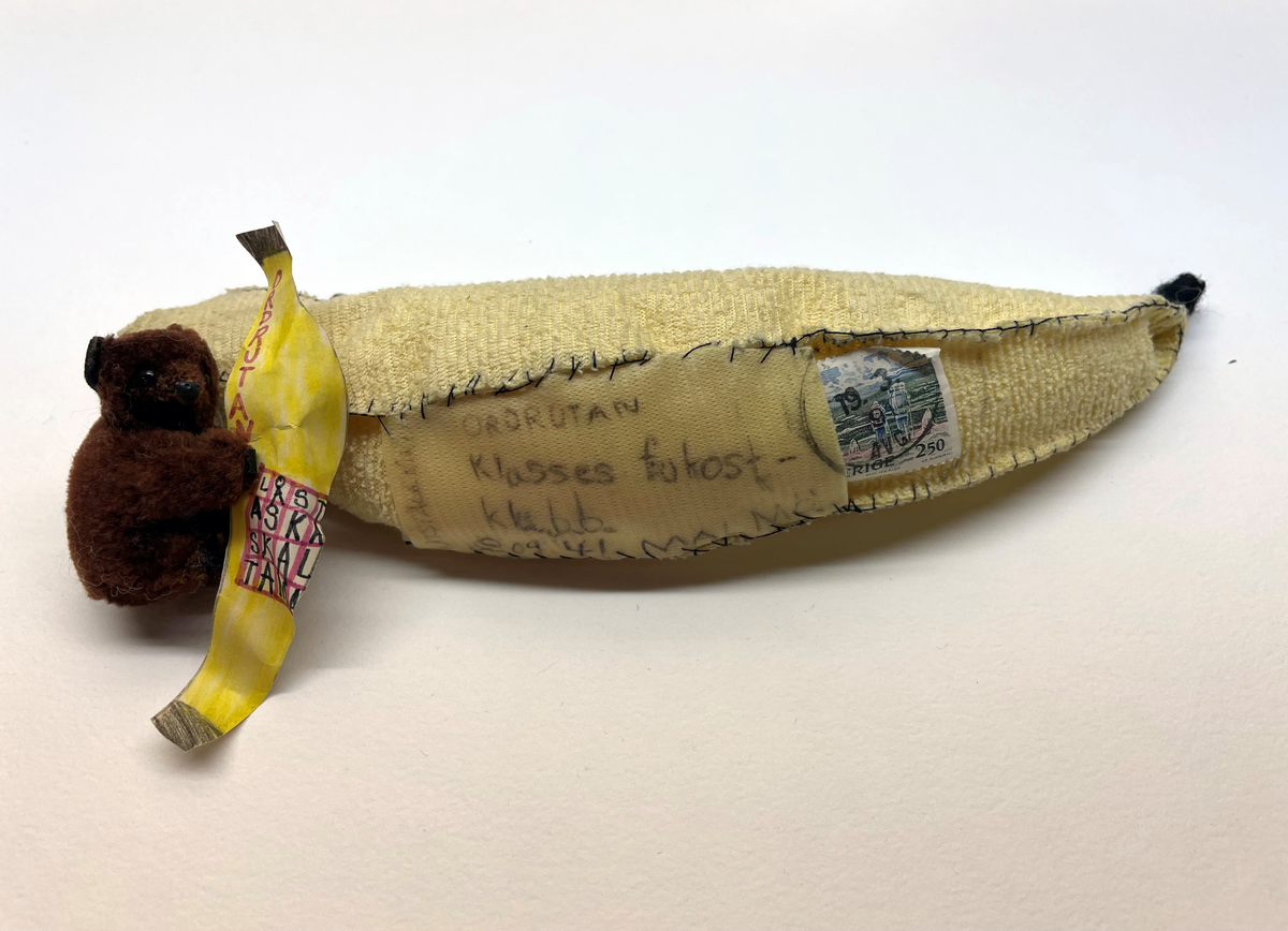 Banan med dragkedja (pennfack?) Bananen är sydd i tyg och ser hemmagjord ut. I bananen finns en brun leksaksbjörn som håller i en utklippt färglagd pappersbanan med svaret på en rebus / ordfläta / korsort ritat. På pappersbananens baksida finns avsändarens namn och adress.

Bananen är adresserad till Klasses frukostklubb, frankerad med ett frimärke (fasttejpade) och poststämplat 1991.
