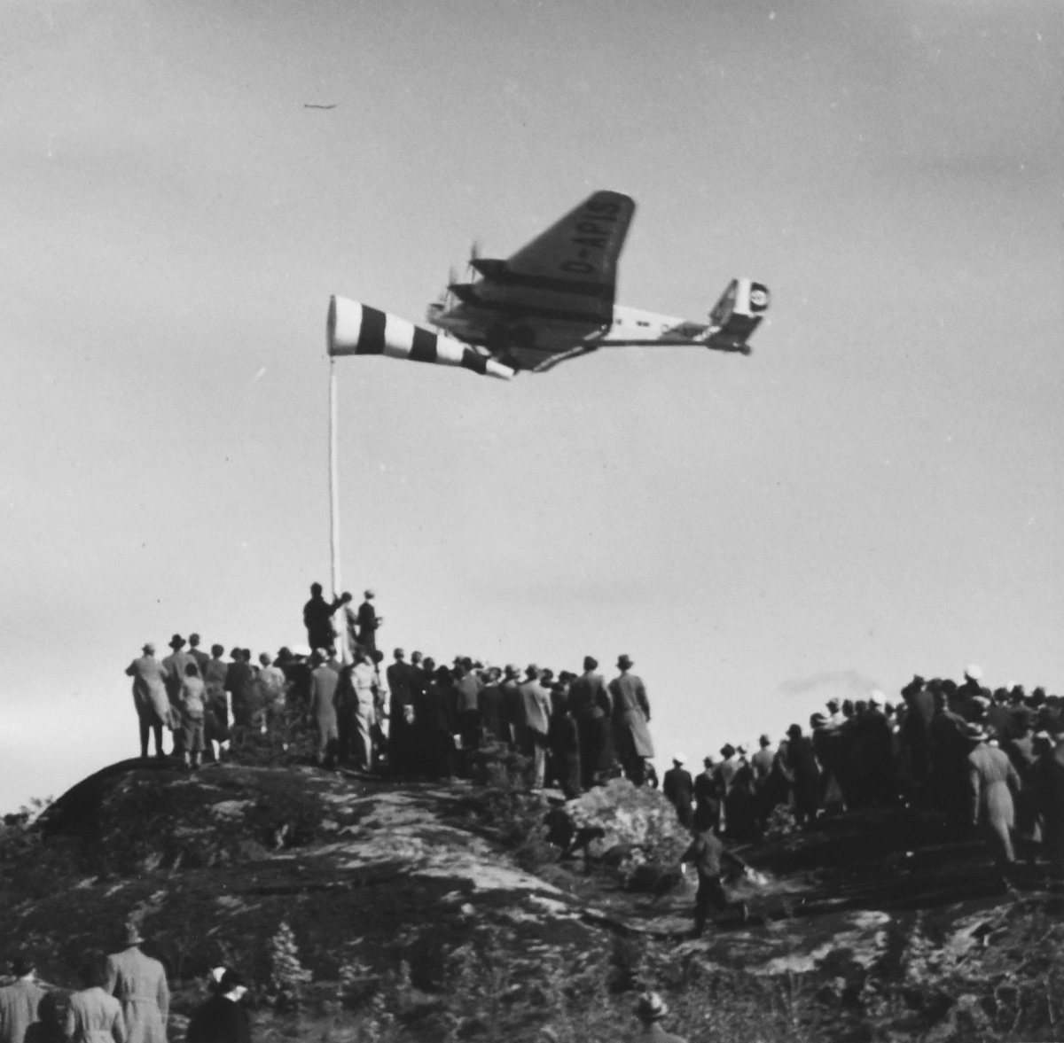 Besökare står på en berghäll för att se ett transportflygplan i luften under invigningen av Bromma flygplats den 23 maj 1936.

Text vid foto: "Invigningen av Bromma"