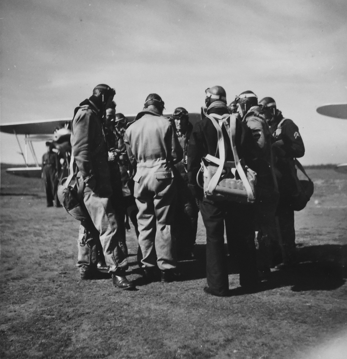 Militära flygare samlas på flygfältet inför flygning, omkring 1934-36. Nio flygare klädda i flygdräkt står i en ring. I bakgrunden står flygtekniker och flygplan.

Text vid foto: "Lärarna till mej"
