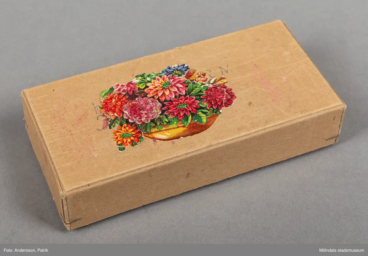 En brunfärgad avlång rektangulär ask som innehåller vaxkritor. På askens ovansida finns ett bokmärke påklistrat i mitten. Bokmärket föreställer en skål med röda och oranga blommor och gröna blad. Asken har tillhört Karin Hansson och hon använde dem när hon var 8-9 år gammal.