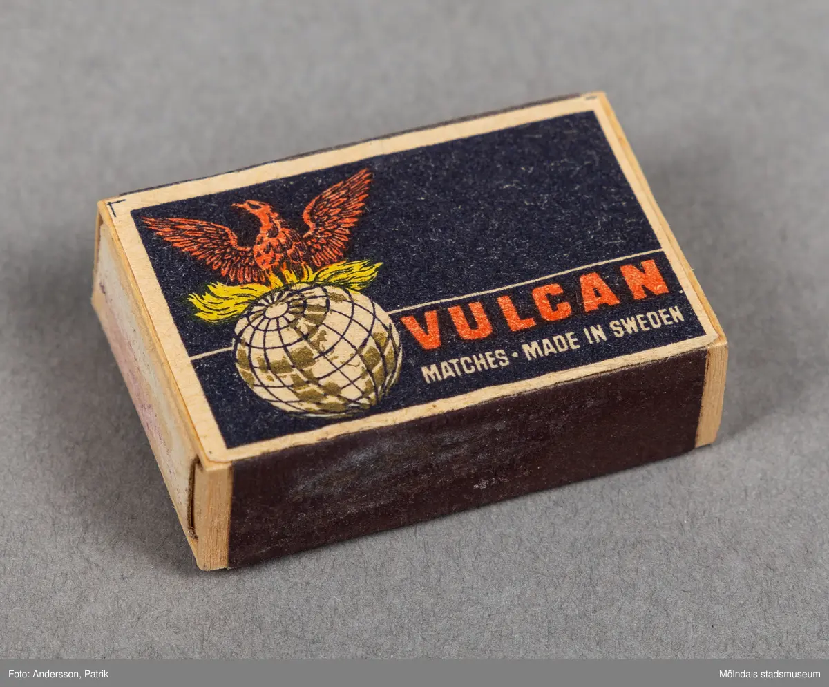 En liten tändsticksask av märket Vulkan. Asken är svart med cremefärgad ram i ytterkanten. På asken finns en bild av en jordglob varpå en röd fågel sträcker ut vingarna. Vulcans tändsticksfabrik grundades av Von Essen släkten 1868. Omkring 1900 var Vulcan världens största tändsticksproducent. År 1903 slog fabriken Vulcan ihop med Jönköpings tändsticksfabrik, Annebergs tändsticksfabrik till Jönköpings och Vulcans tändsticksfabrik.