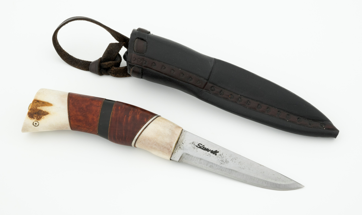 Tollekniv i slire, der knivskaftet og slira er lagd av den danske etnologen, museumslederen og knivmakeren Henrik Vensild, som i mange år besøkte Norsk skogmuseum og det såkalte «knivtorget» under De nordiske jakt- og fiskedager.

Kniven er 20,0 centimeter lang, målt fra bladspissen til og med knappen i ytterenden av skaftet. Bladet er 9,1 centimeter langt og snaut 2,3 centimeter bredt ved brystningen (overgangen til skaftet). Her er bladtjukkelsen om lag 3,7 millimeter, men den avtar noe framover mot knivspissen. Umiddelbart bak slipefasen der er tjukkelsen snaut 2,6 millimeter. Overflata på bladet har den noe rue overflata den fikk av hammeren på ambolten intakt, men disse flatene er polerte. Bakerst på høyre side finner vi et stempel, «Steen N.», som er stempelet til den danskfødte smeden Steen Nielsen, som lenge har bodd i Kristiansand. Bladryggen er planslipt, men er samtidig noe avrundet i den fremre enden. Slipefasene er opptil 6,5 millimeter brede.

Skaftet er nesten 10,5 centimeter langt. Det buer noe nedover (mot skaftbuken) i den bakre enden og har, som knivblader flest, et ovalt tverrsnitt. Sett i lengderetningen er den midtre delen av denne knivdelen noe konveks. Skaftet er sammensatt av flere komponenter. Fremst, mot bladet, er det en del av horn, antakelig reinsdyrgevir. Den enden som vender mot bladet er rett (butt), den andre enden er skrått avskåret. Mot denne flata er det lagt ei lærforing, deretter ei sølvplate og så nok ei lærforing. Deretter følger et stykke brunt, mønstret tre, en svart ring av et ibenholtliknende materiale. og nok et stykke av det brune, mønstrete treet. Ytterenden er skråskåret. På den skrå endeflata er det lagt nok ei lærforing, denne gangen uten noe metallinnlegg, mot endetappen, som er av reisdyrhorn. På denne komponenten er det to små ringer med et punkt i sentrum, en slags signatur for Henrik Vensild. Tangen fra knivbladet er ført gjennom hele skaftet. Den bakre enden av denne jernteinen er klinket mot noe som later til å være en messingring.

Slira er sydd av mørkebrunt lær. Den er cirka 16,9 centimeter lang (uten beltestropp) og 3,4 centimeter bred (målt på midten). Slireformen er symmetrisk, når man ser bort fra at overkanten er skrått avskåret. Den nedre delen, som skal omslutte bladet, er nokså flattrykt, mens den øvre delen, som skal omslutte nedre del av skaftet, er mer rundoval. Sliras forside er dekorert med to parallelle linjer i lengderetningen med innstemplete små treflikete blader med noenlunde regelmessig innbyrdes avstand. Dette dekorelementet er, åpenart med hensikt, plassert på en måte som bryter litt med den nevnte symmetrien. Like under sliremunningen er det skåret små spalter i slirelæret. Her er det inntredd ei cirka 9 millimeter bredt lærreim, som også er tredd gjennom ei spalte i et «øre» på den øvre delen av sliresømmen. Endene på den nevnte lærreima er knyttet sammen slik at den danner ei løkke som kan tres inn på et belte. Henrik Vensild har stemplet inn initialene sine og produksjonsåret - 2005 - på baksida av sliras overdel.