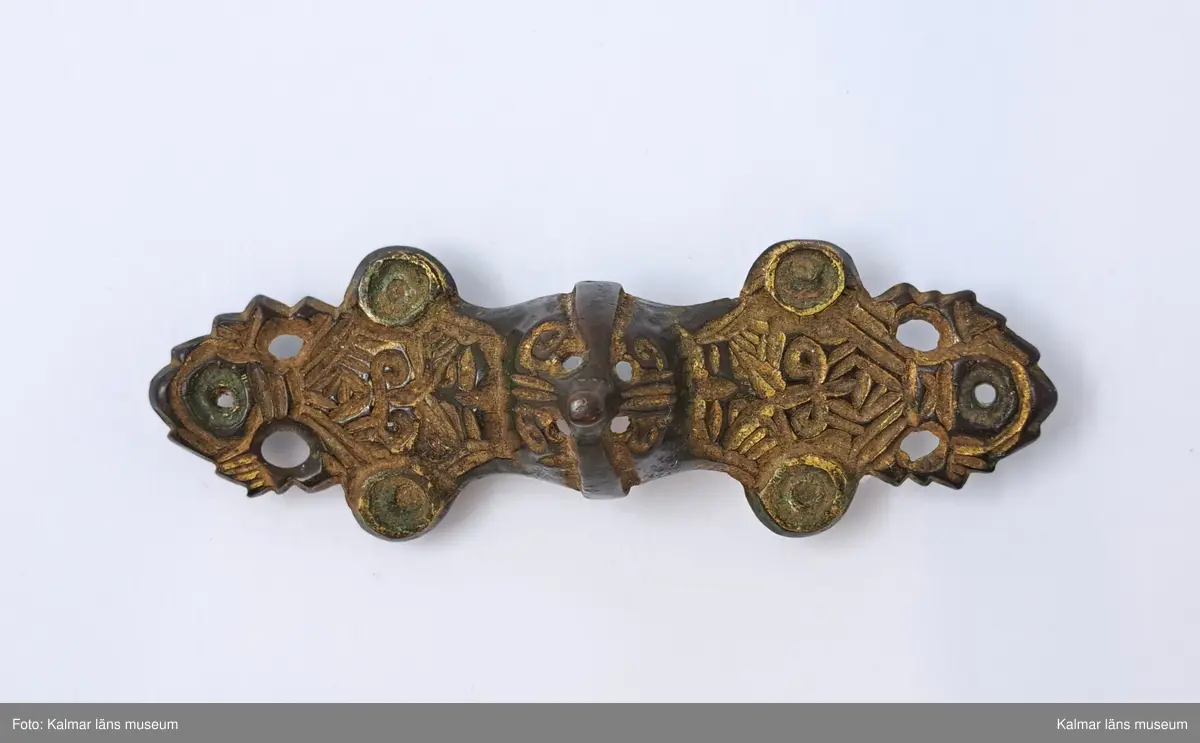 KLM 6. Spänne, likarmat spänne. Gjutet av brons. Mittornamentet gjutet i ett stycke med spännet. Vardera ändstycket har tre utstående flikar med rundlar, alla genomborrade. Spår av förgyllning. På undersidan finns nålfäste och nålhållare kvar men själva nålen saknas.