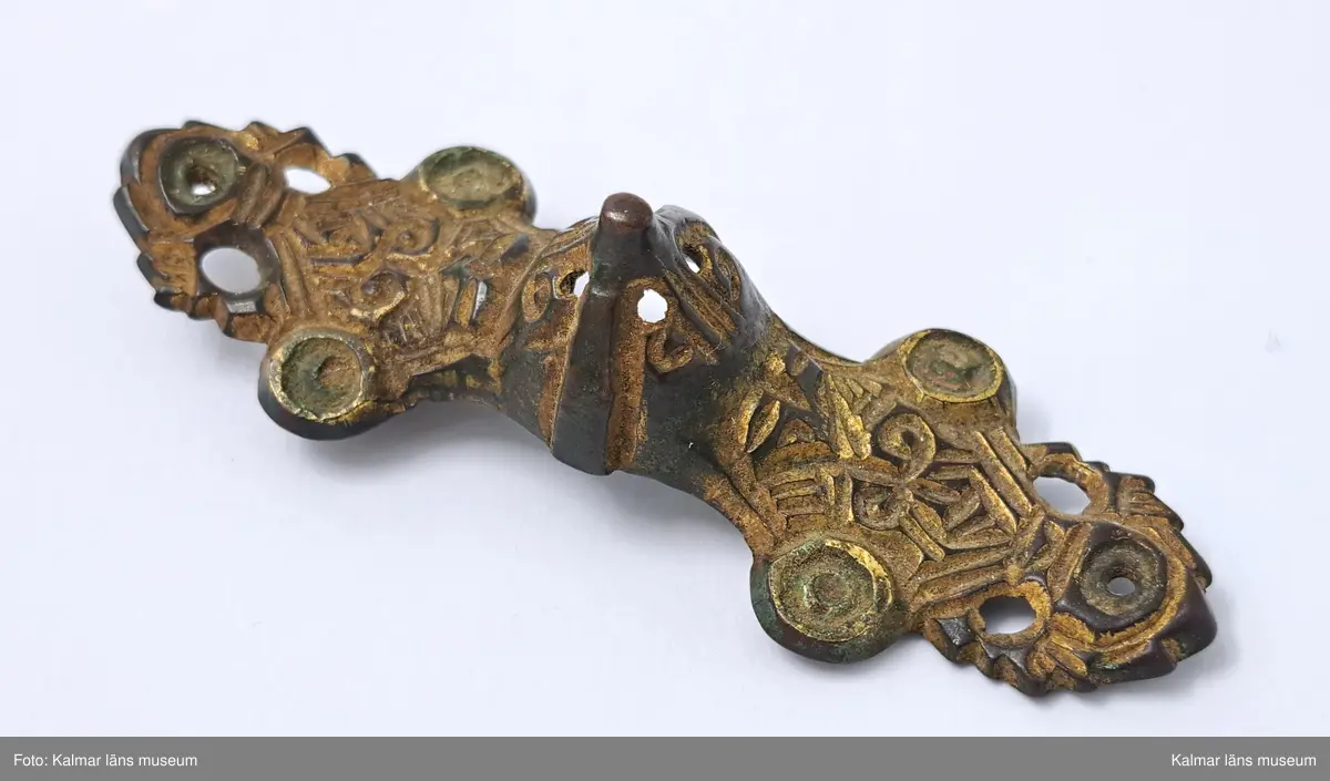 KLM 6. Spänne, likarmat spänne. Gjutet av brons. Mittornamentet gjutet i ett stycke med spännet. Vardera ändstycket har tre utstående flikar med rundlar, alla genomborrade. Spår av förgyllning. På undersidan finns nålfäste och nålhållare kvar men själva nålen saknas.