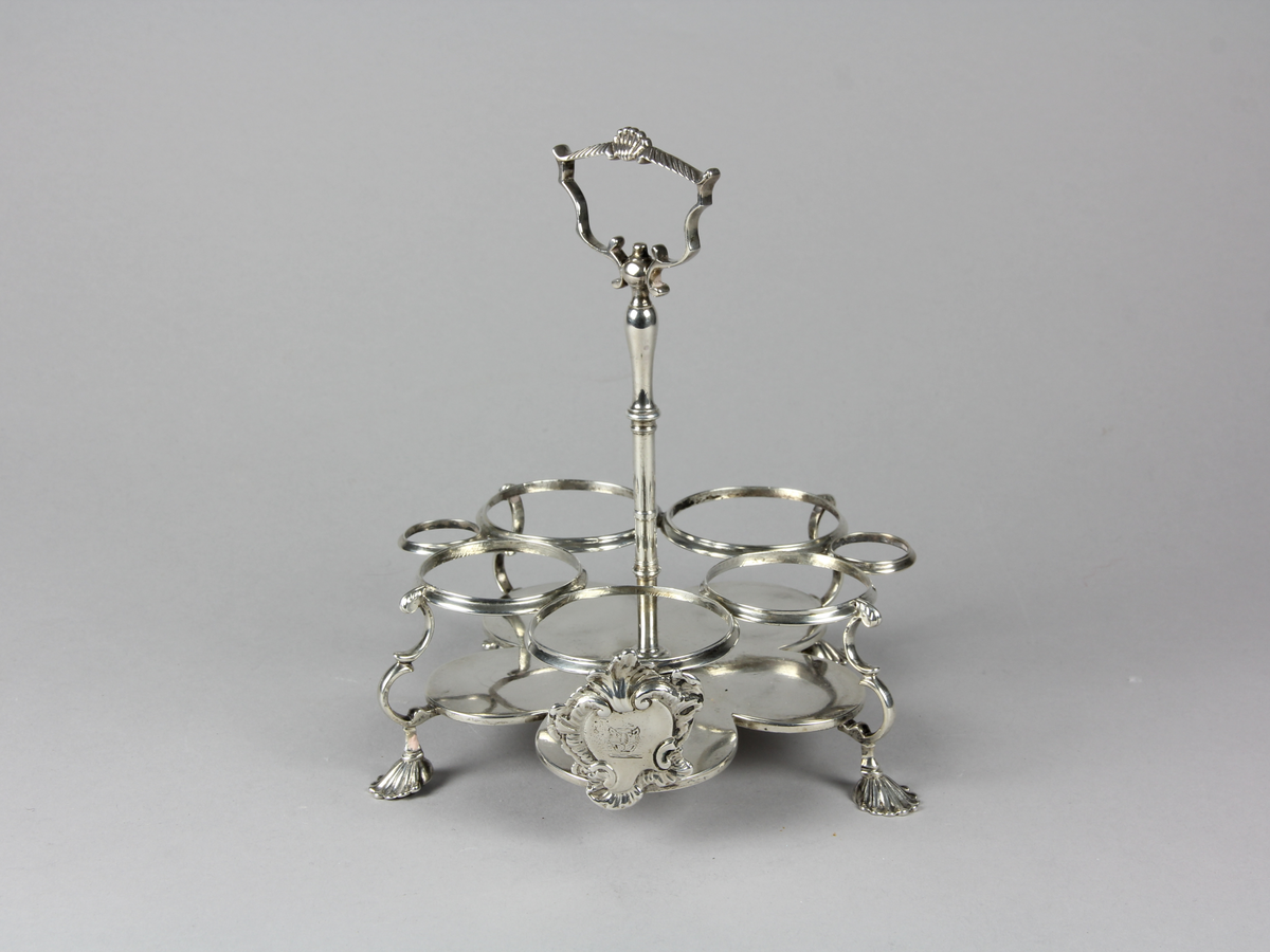 Bordsställ av silver för flaskor och dosor av glas. Ställ med cinquefoil formad platta som stöds av fyra svängda ben med snäckformade fotavslutningar. Övre delen med sju stycken cirkelformade ställ för flaskor och dosor, mitt fram på övre delen en kartusch med oidentifierat familjevapen. I mitten en stång med profilerat handtag som kröns upptill med en mussla.