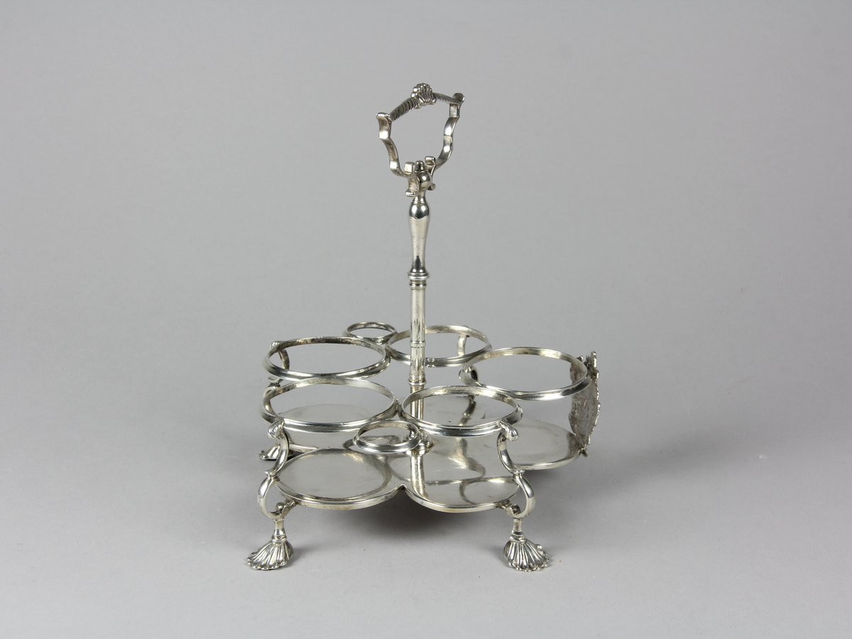 Bordsställ av silver för flaskor och dosor av glas. Ställ med cinquefoil formad platta som stöds av fyra svängda ben med snäckformade fotavslutningar. Övre delen med sju stycken cirkelformade ställ för flaskor och dosor, mitt fram på övre delen en kartusch med oidentifierat familjevapen. I mitten en stång med profilerat handtag som kröns upptill med en mussla.