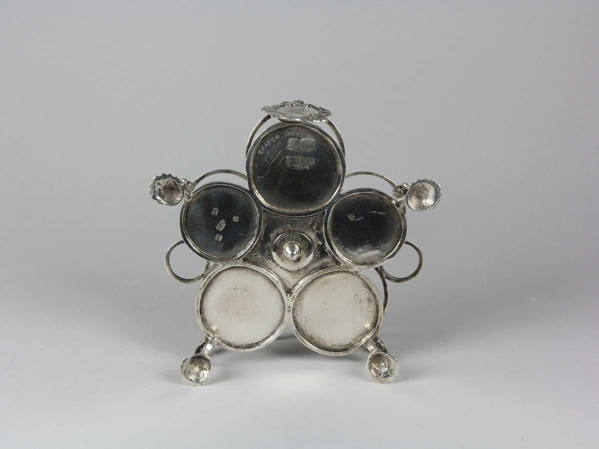 Bordsställ av silver för flaskor och dosor av glas. Ställ med cinquefoil formad platta som stöds av fyra svängda ben med snäckformade fotavslutningar. Övre delen med sju stycken cirkelformade ställ för flaskor och dosor, mitt fram på övre delen en kartusch med oidentifierat familjevapen. I mitten en stång med profilerat handtag som kröns upptill med en mussla.