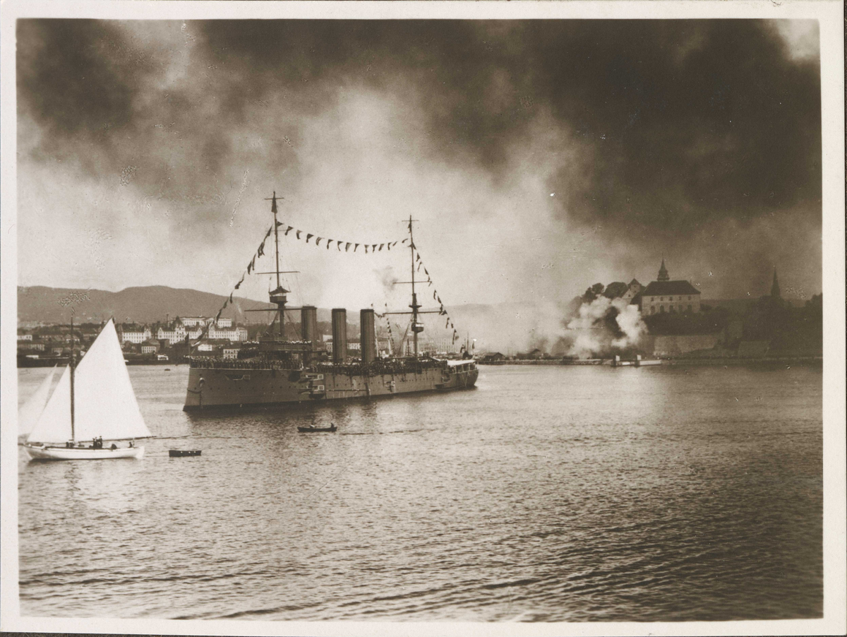 I forgrunnen Oslofjorden og HMS Cornwall som hilser KRISTIANIAFJORD med stor flagging og mannet reling. I bakgrunnen Akershus festning i røyksky etter salutt. Mindre seilbåt t.v.