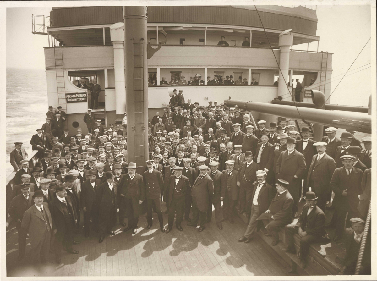 Gruppefotografering av passasjerer. Kong Haakon i midten foran med Jørgen Løvland til venstre. F. v. i første rekke: Johan Castberg og Thomas Cathinco Bang. Kristofer Lehmkuhl nr. 4 f.h. på balkongen bak.