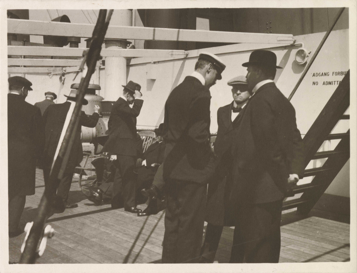 Passasjerer i samtale på promenadedekket. F.h.: Gustav Henriksen, Hans Jørgen Darre-Jenssen, Kong Haakon. I bakgrunnen Thomas Cathinco Bang som hytter med neven.
