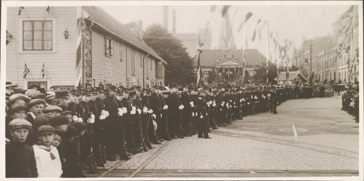 Folkemengde og soldater i Rådhusgata avventer Kongens besøk med KRISTIANIAFJORD. Kristiansand Børs og Kristiansand domkirke i bakgrunnen.