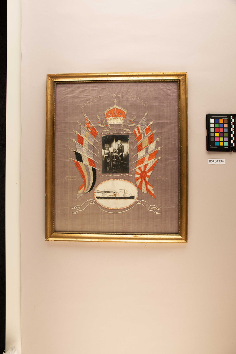 Brodert silkebilde av DS ERLE i stående format. Midt i motivet fotografi av ukjent sjømann til rors, omkranset av seks ulike flagg på hver side og krone øverst. Under et brodert bilde av DS ERLE under fart..
