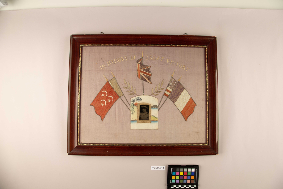 Brodert silkebilde med fotografi av ung mann. Midt i motivet fem ulike flagg som dekor rundt fotografiet. Fransk flagg, amerikansk flagg, Storbritannias flagg og trolig Ottomansk flagg (halvmåne og stjerner).