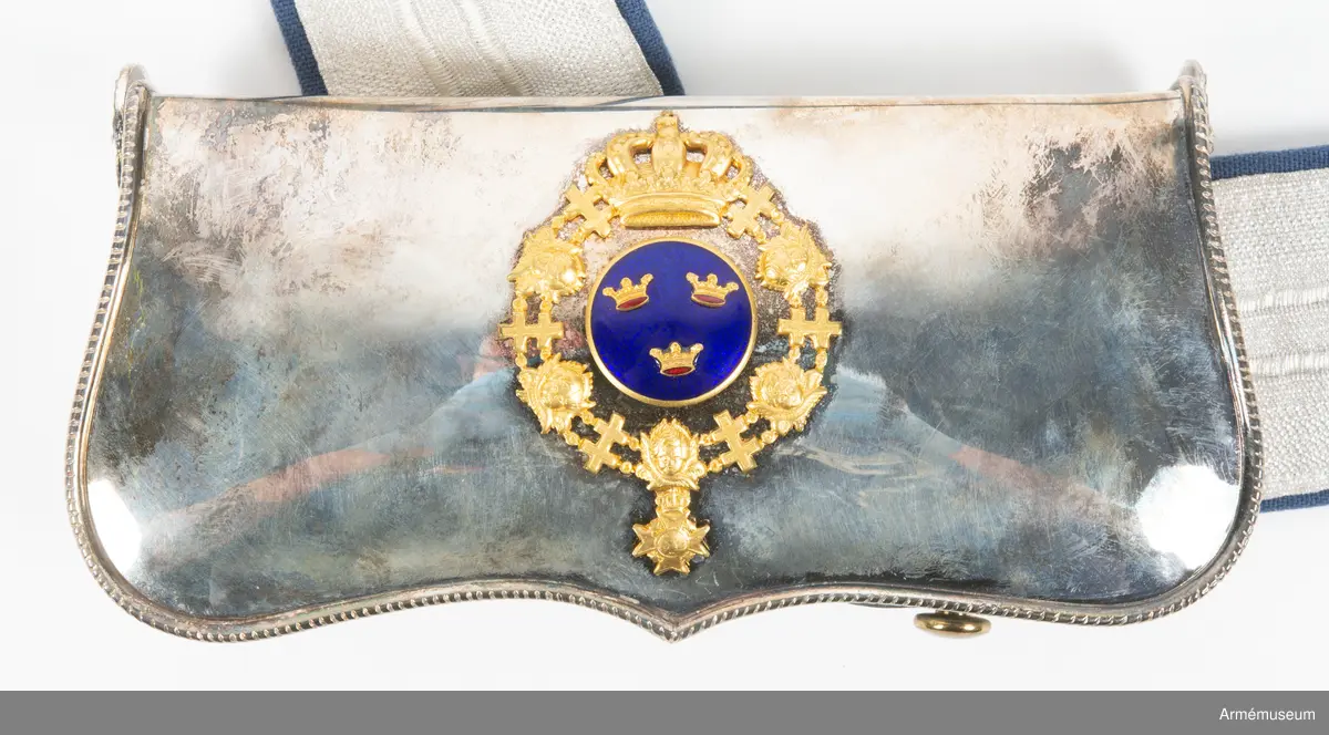 Kartuschlåda av silver med tillhörande kartuschrem.

Lådan är gjord av mässing medan locket och kortsidorna är av silver. Fram- och baksidan av lådan är klädd med ljusbrunt skinn. Locket är dekorerat med Serafimerordens krönta och förgyllda ordenskrans kring en emaljerad knapp med tre förgyllda kronor i mitten. Dekorationen är gjuten separat och fäst på lockets baksida med vikben och en stor skruv. Lådans kortsidor pryds upptill av små lejonmaskaroner i silver.
På baksidan av lådan sitter ett intrikat system av läderremmar för att spänna fast kartuschremmen som är tudelad och gjord av 5,1 cm bred silvergalon, fodrad med blått bomullstyg. Samtliga spännen på remmen är av silver.
Både låda och spännen är märkta med silverstämplar: KLC, (kattfot?) och O10.