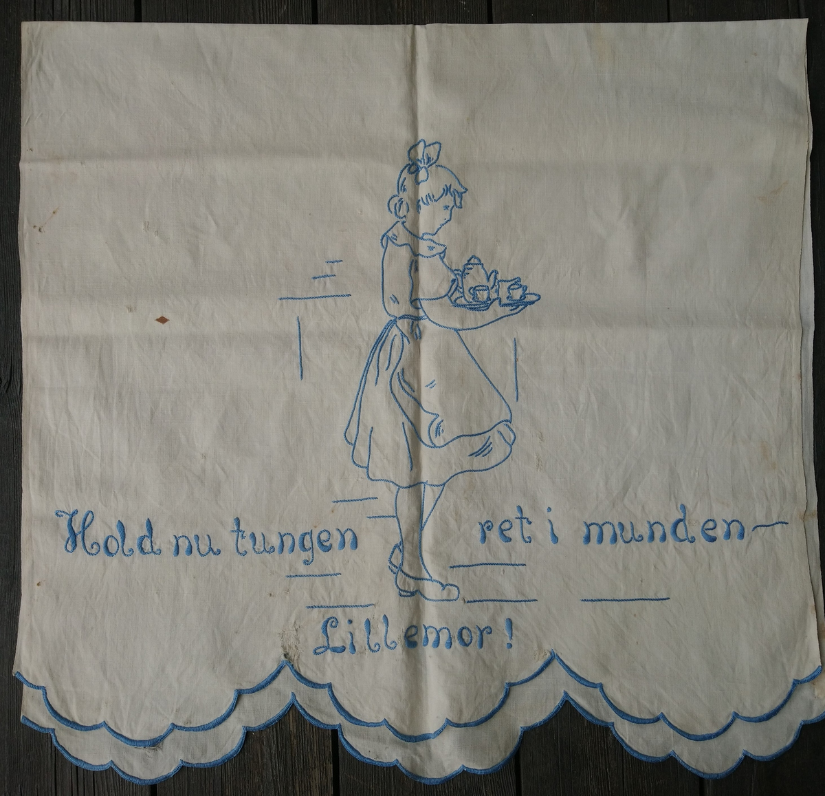 Hvitt håndkle med blått broderi. Motivet er ei jente som bærer et serveringsbrett og teksten "Hold nu tungen ret i munden Lillemor!"