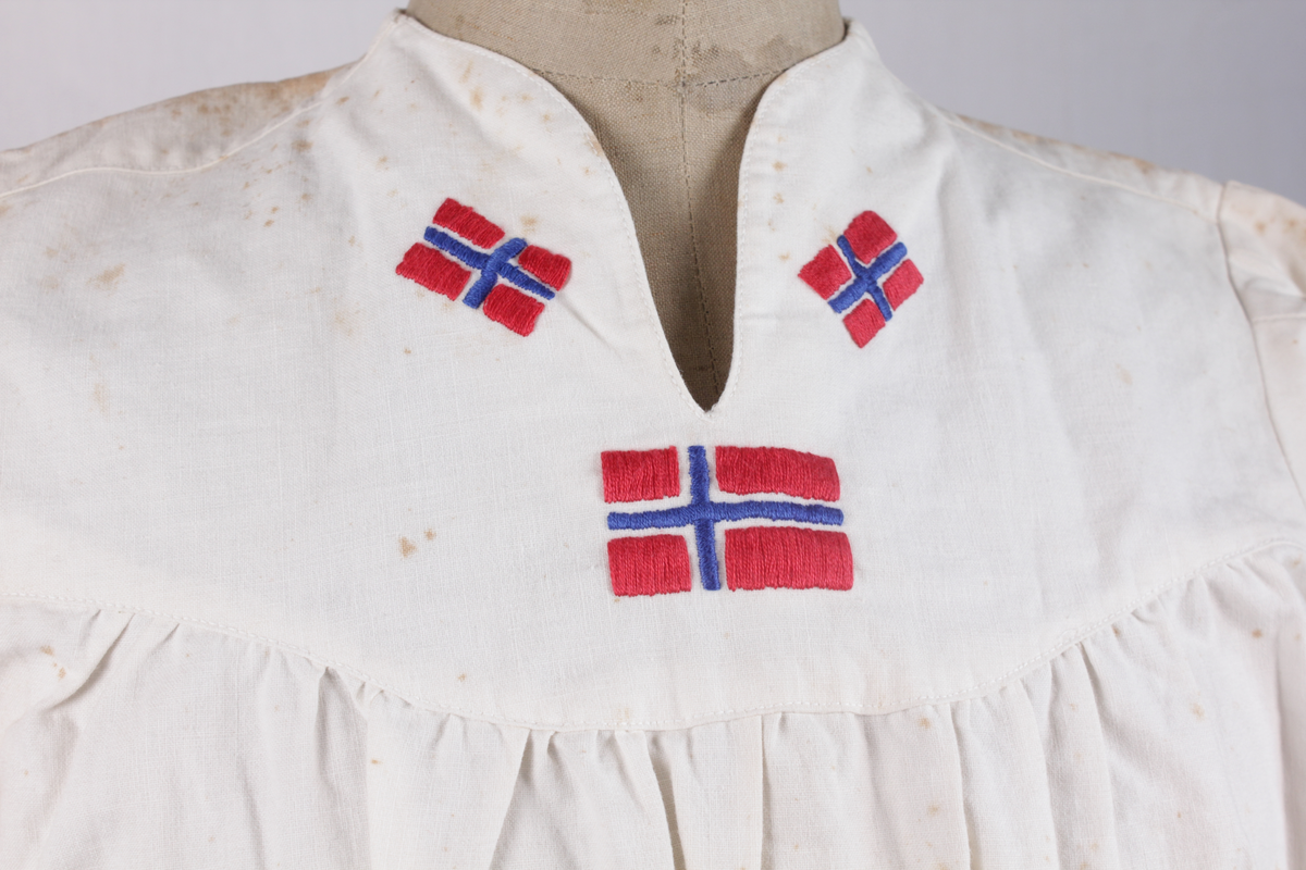 Hvit 17. mai-kjole fra 1945. Sydd av blekede sukkersekker eller melsekker og brodert med norske flagg. Kjolen har 12 falske knapper på rygg og lukkes med seks skjulte trykknapper. Glidelås i siden.