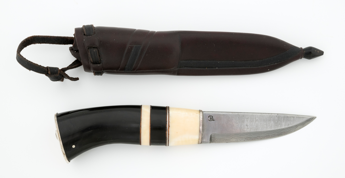 Tollekniv i slire, der knivskaftet og slira er lagd av den danske etnologen, museumslederen og knivmakeren Henrik Vensild, som i mange år besøkte Norsk skogmuseum og det såkalte «knivtorget» under De nordiske jakt- og fiskedager.

Kniven er 20,0 centimeter lang, målt fra bladspissen til og med knappen i ytterenden av skaftet. Bladet, som er damasksmidd, er 8,8 centimeter langt og 2,5 centimeter bredt ved brystningen (overgangen til skaftet). Her er bladtjukkelsen om lag 3,7 millimeter, men den avtar noe framover mot knivspissen. Umiddelbart bak slipefasen der er tjukkelsen 2,6 millimeter. Overflata på bladet var glattslipt og polert. Bakerst på høyre side finner vi et stempel, «B», som er stempelet til smeden. Bladryggen er rettslipt i lengderetningen og noe avrundet i tverrsnitt. Den fremre enden skrår noe nedover. Slipefasene er opptil 7,5 millimeter brede.

Skaftet er nesten 11,2 centimeter langt. Det buer noe nedover (mot skaftbuken) i den bakre enden og har, som knivblader flest, et ovalt tverrsnitt. Sett i lengderetningen er den fremre halvdelen av denne knivdelen noe konveks. Skaftet er sammensatt av flere komponenter. Fremst, mot bladet, er det ei 2,5 millimeter bred metallskive, muligens av nysølv. Deretter følger en bit av gulkvitt hornmateriale, ei nysølvskive, så en noe bredere bit av et svart, ibenholtliknende materiale. Så følger ei skive av dset kvite hornmaterialet, og deretter en stor bit av det svarte ibenholtliknende materialet. På den bakre delen er det inntappet to sølvstifter, antakelig som et slags varemerke for knivmakeren Henrik Vensild. I den bakre enden, som er skråskåret, er det nok ei drøyt 2 millimeter tjukk nysølvplate. Mellom de nevnte skaftdelene ligger det foringer av lær. Tangen fra knivbladet er ført gjennom hele skaftet. Den bakre enden av denne jernteinen er klinket mot noe som later til å være en messingring.

Slira er sydd av mørkebrunt lær. Den er cirka 19,9 centimeter lang (uten beltestropp, men målt ved enden av sømmen på baksida) og 3,6 centimeter bred (målt på midten). Slireformen er symmetrisk, når man ser bort fra at overkanten er skrått avskåret. Den nedre delen, som skal omslutte bladet, er nokså flattrykt, mens den øvre delen, som skal omslutte nedre del av skaftet, er mer rundoval. Sliras forside er dekorert med linjer i lengderetningen og noe skrått på tvers av overdelen. Dekorelementene er, åpenbart med hensikt, plassert på en måte som bryter litt med den nevnte symmetrien i sliras ytre form. Like under sliremunningen er det skåret små spalter i slirelæret. Her er det inntredd ei drøt 7,5 millimeter bredt lærreim, som også er tredd gjennom ei spalte i et «øre» på den øvre delen av sliresømmen. Endene på den nevnte lærreima er knyttet sammen slik at den danner ei løkke som kan tres inn på et belte. I den nedre enden av slira er det innsatt en knapp av et ibenholtliknende materiale. Henrik Vensild har stemplet inn initialene sine og og sifrene «04» - som antakelig forteller at produksjonsåret var 2004 - på baksida av sliras overdel.