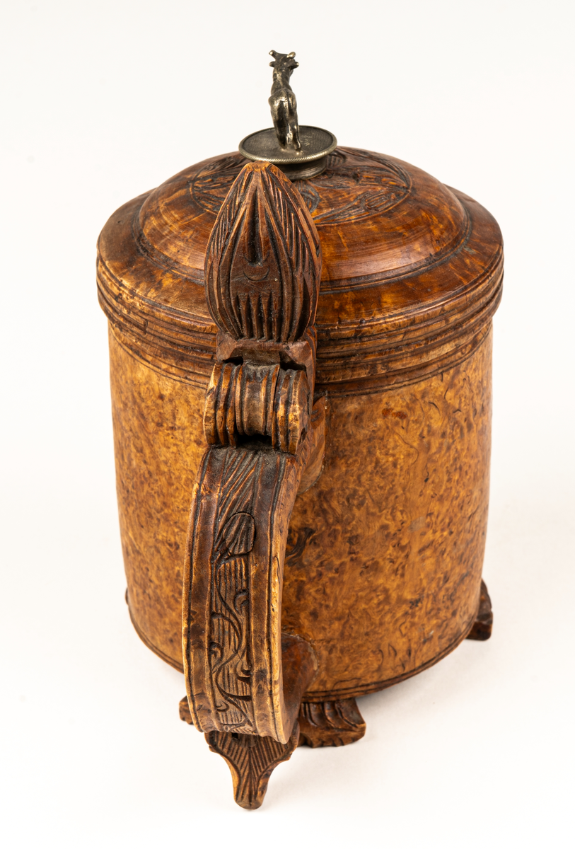 Dryckeskanna, stånka,  av masur, rundsvarvad ur ett stycke, tre låga förrer formade som blad. Knopp på handtagets övre del. På locket ristade ornament samt tjurfigur i bly. Kannan inuti plåtklädd.