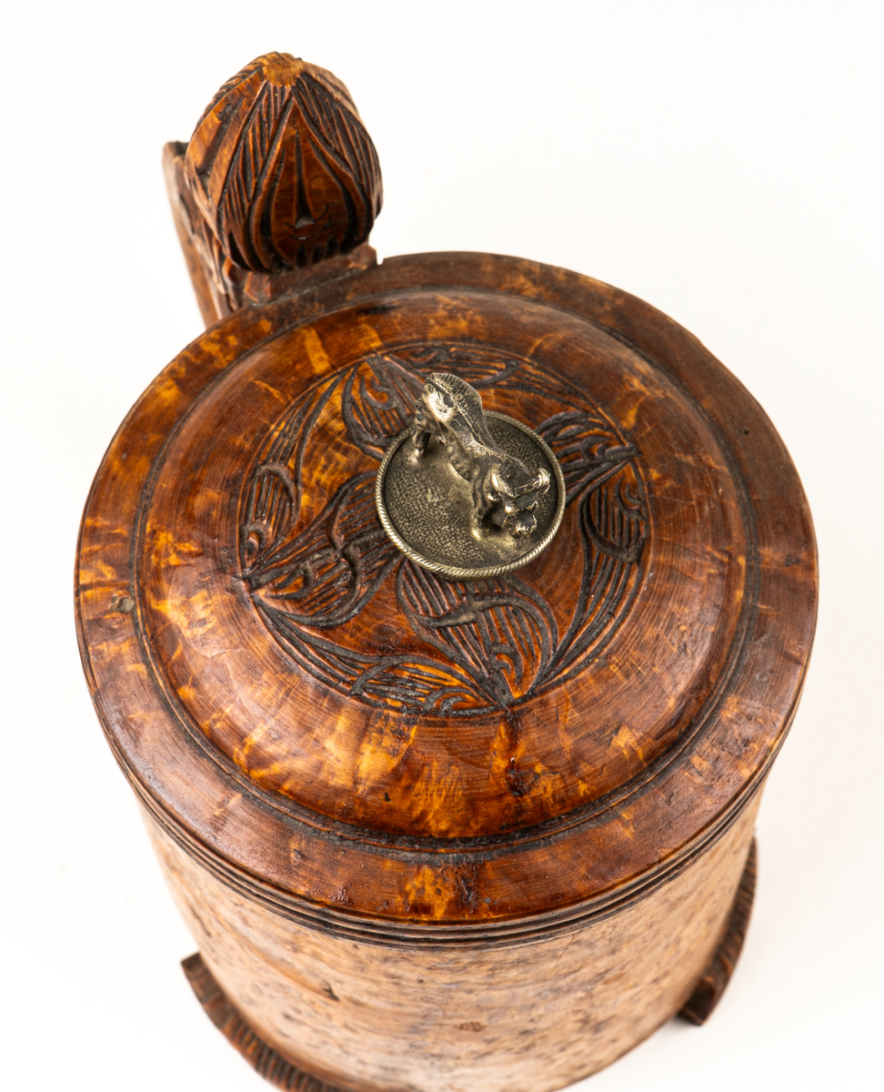 Dryckeskanna, stånka,  av masur, rundsvarvad ur ett stycke, tre låga förrer formade som blad. Knopp på handtagets övre del. På locket ristade ornament samt tjurfigur i bly. Kannan inuti plåtklädd.